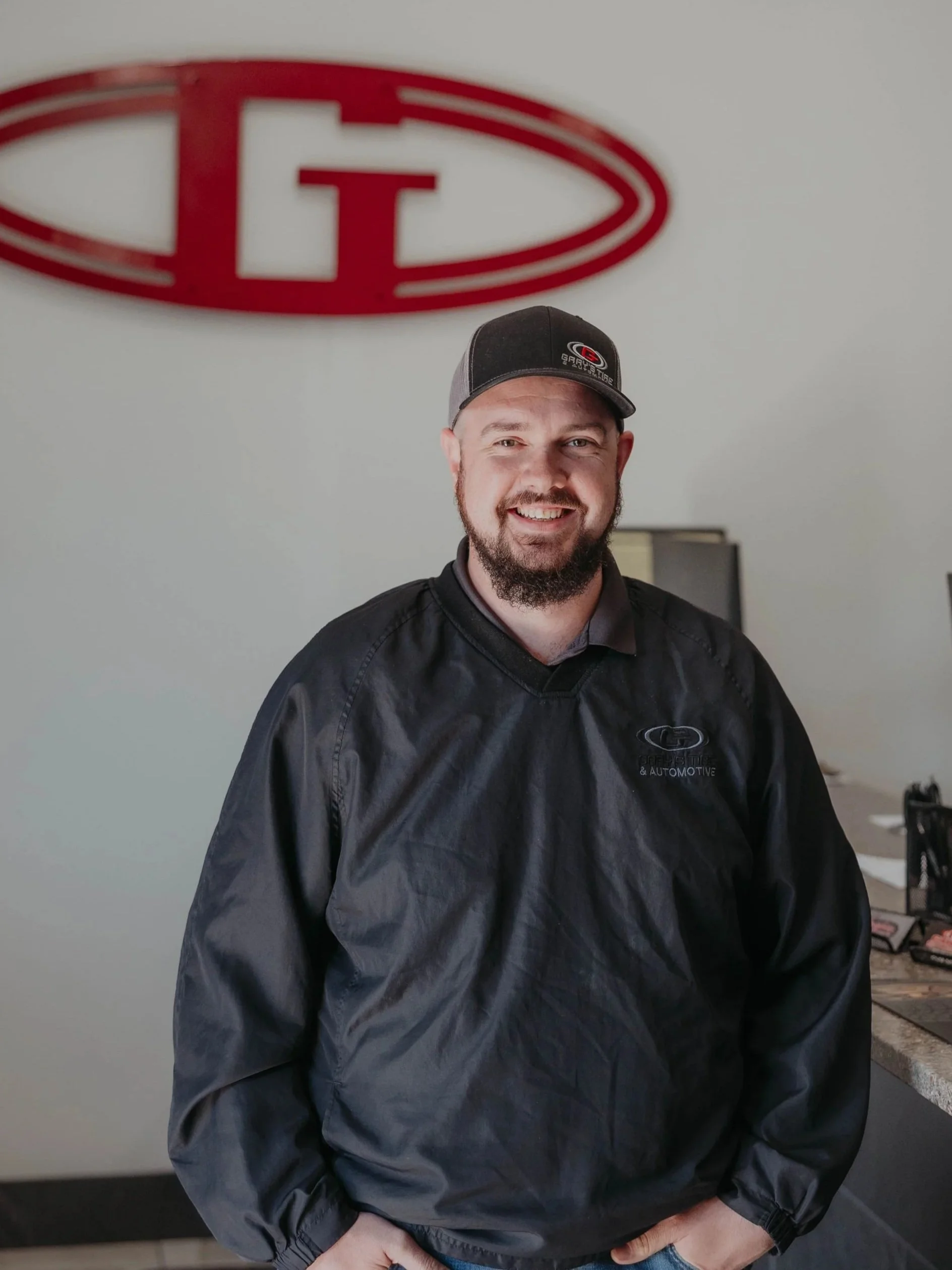 A smiling man with a beard wearing a black baseball cap and black jacket standing in front of a gray wall with a red and white logo above his head.