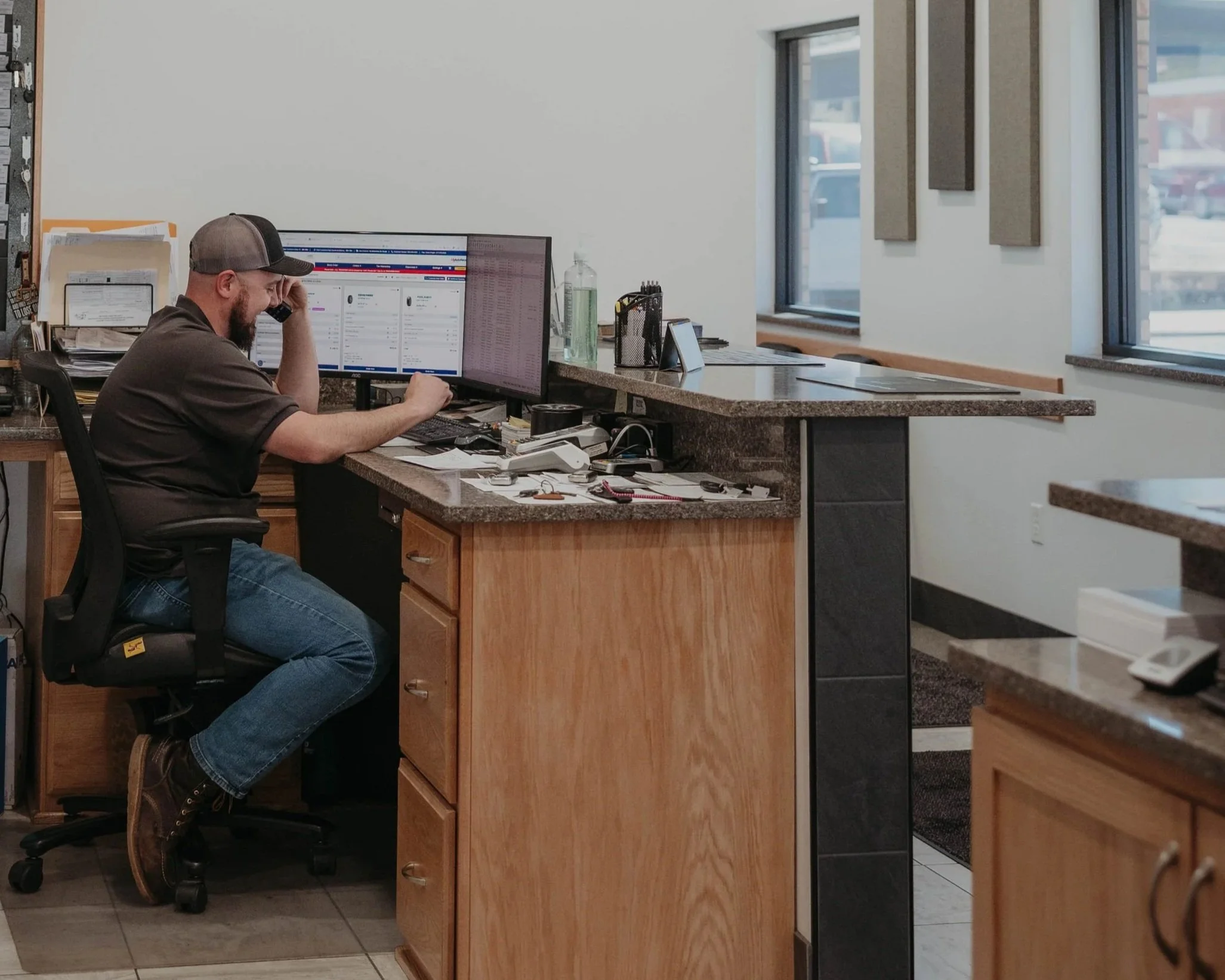 A man with a beard and a baseball cap sitting at a desk in an office, talking on a mobile phone with multiple computer monitors, office supplies, and papers.
