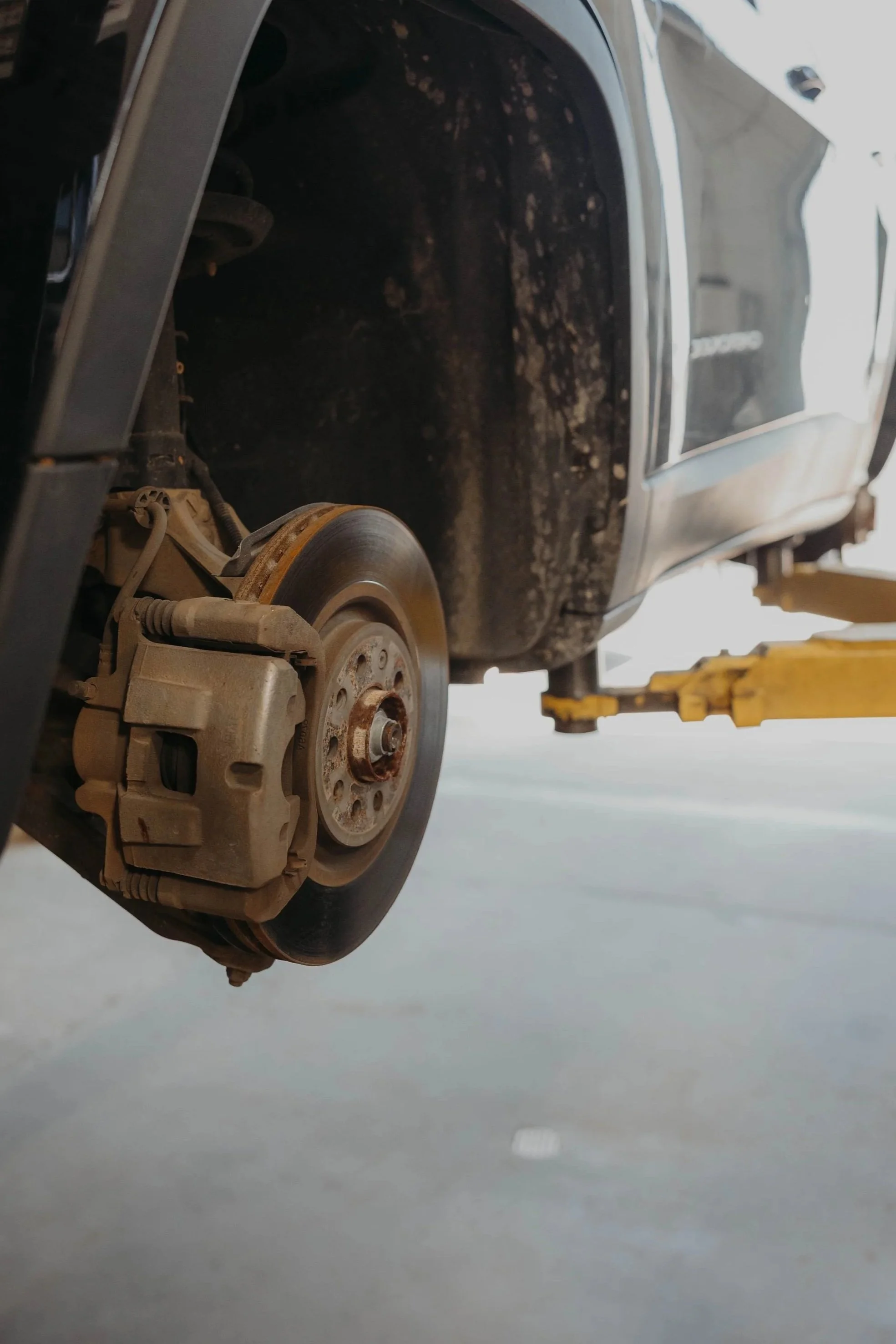 Close-up image of a car's front wheel removed, showing disc brake and suspension components, with the car lifted on a yellow hydraulic lift in a garage or workshop.