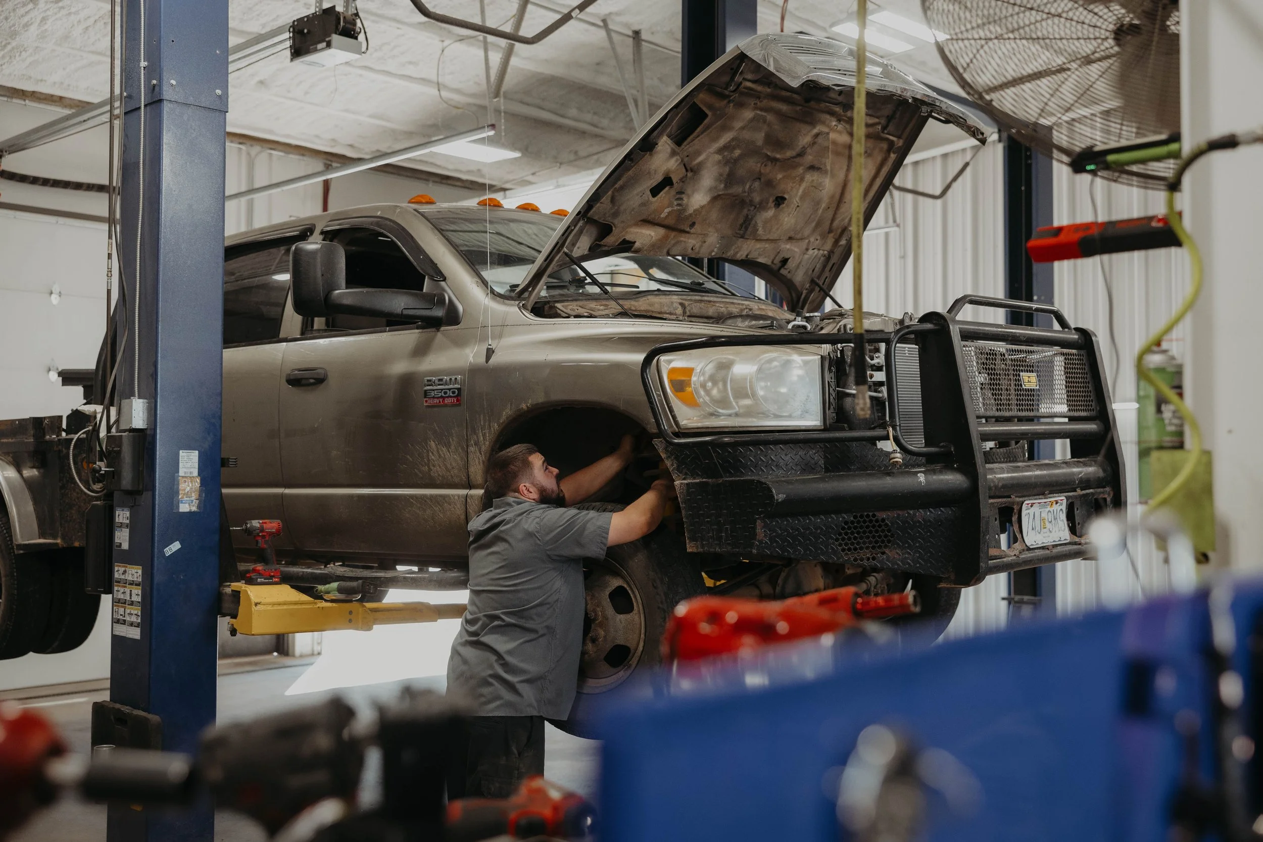 A mechanic working underneath a raised pickup truck with its hood open in an automotive repair shop.