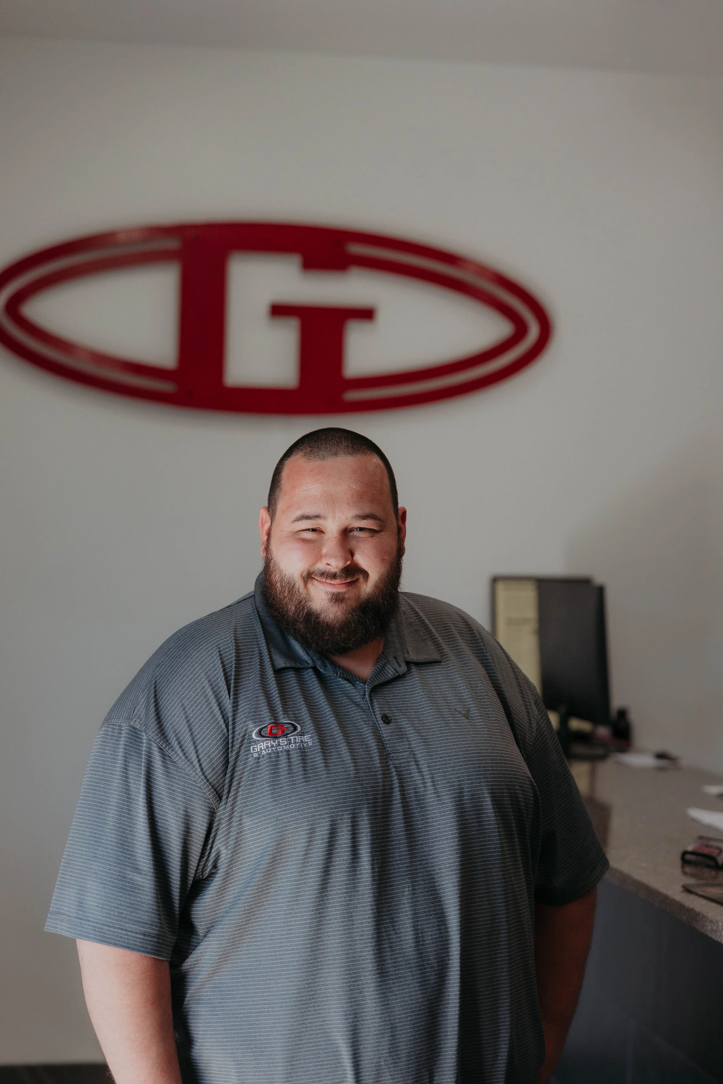 A smiling man with a beard, wearing a gray sale polo shirt with a GARY'S TIRE & AUTOMOTIVE logo, standing in a room with a white wall, a computer monitor, and a framed certificate in the background. A large red and white logo is on the wall above his head.