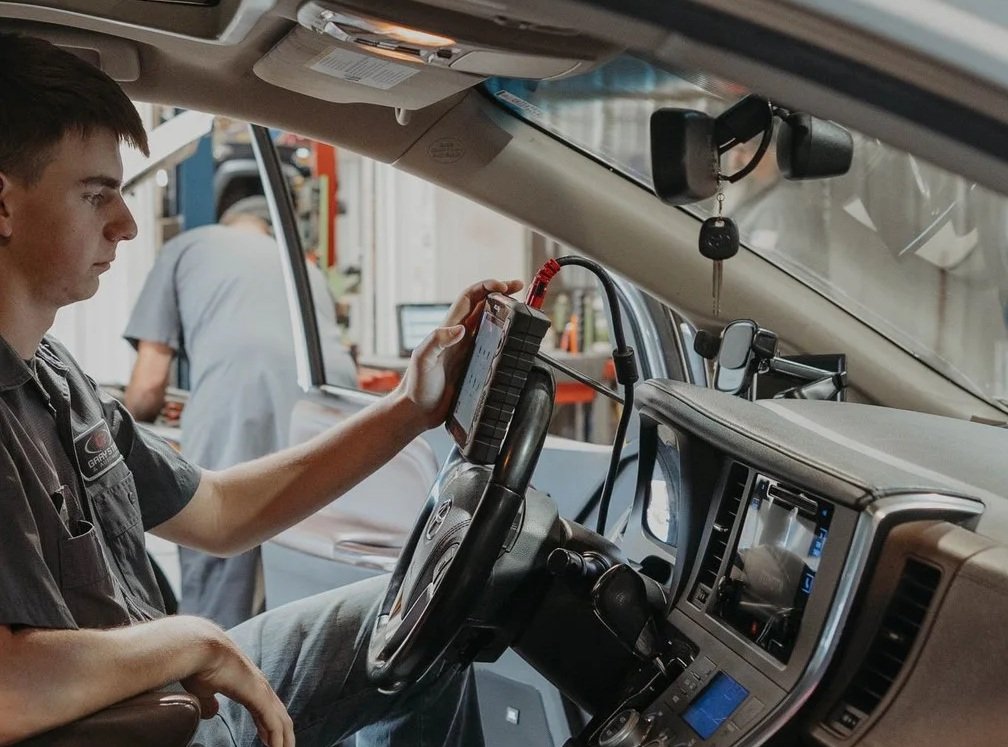 A mechanic sitting in the driver seat of a vehicle, using a diagnostic scanner connected to the vehicle's dashboard during a repair or maintenance task.