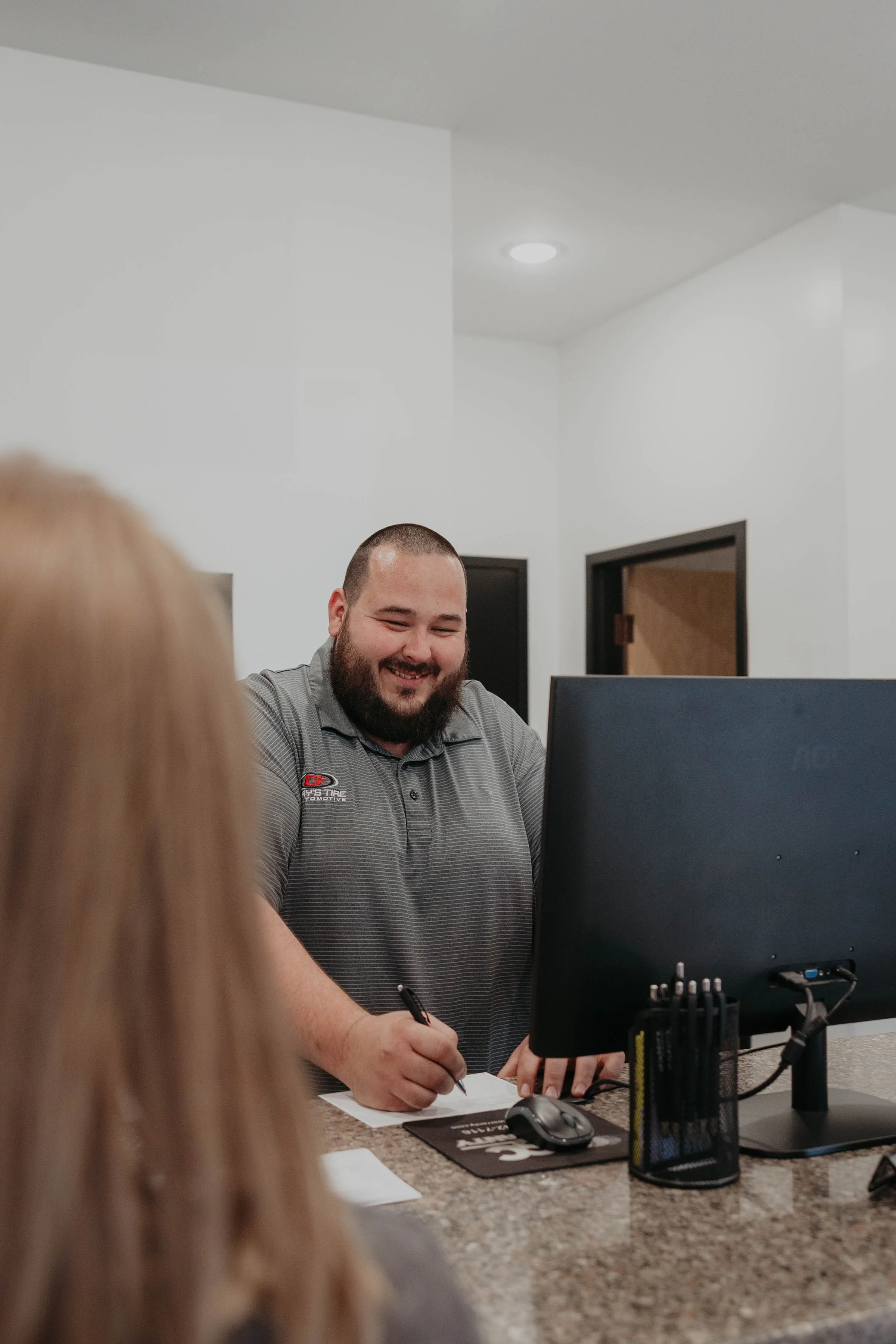 A man with a beard smiling while working at a desk with a computer, paperwork, and office supplies, in a bright office space.