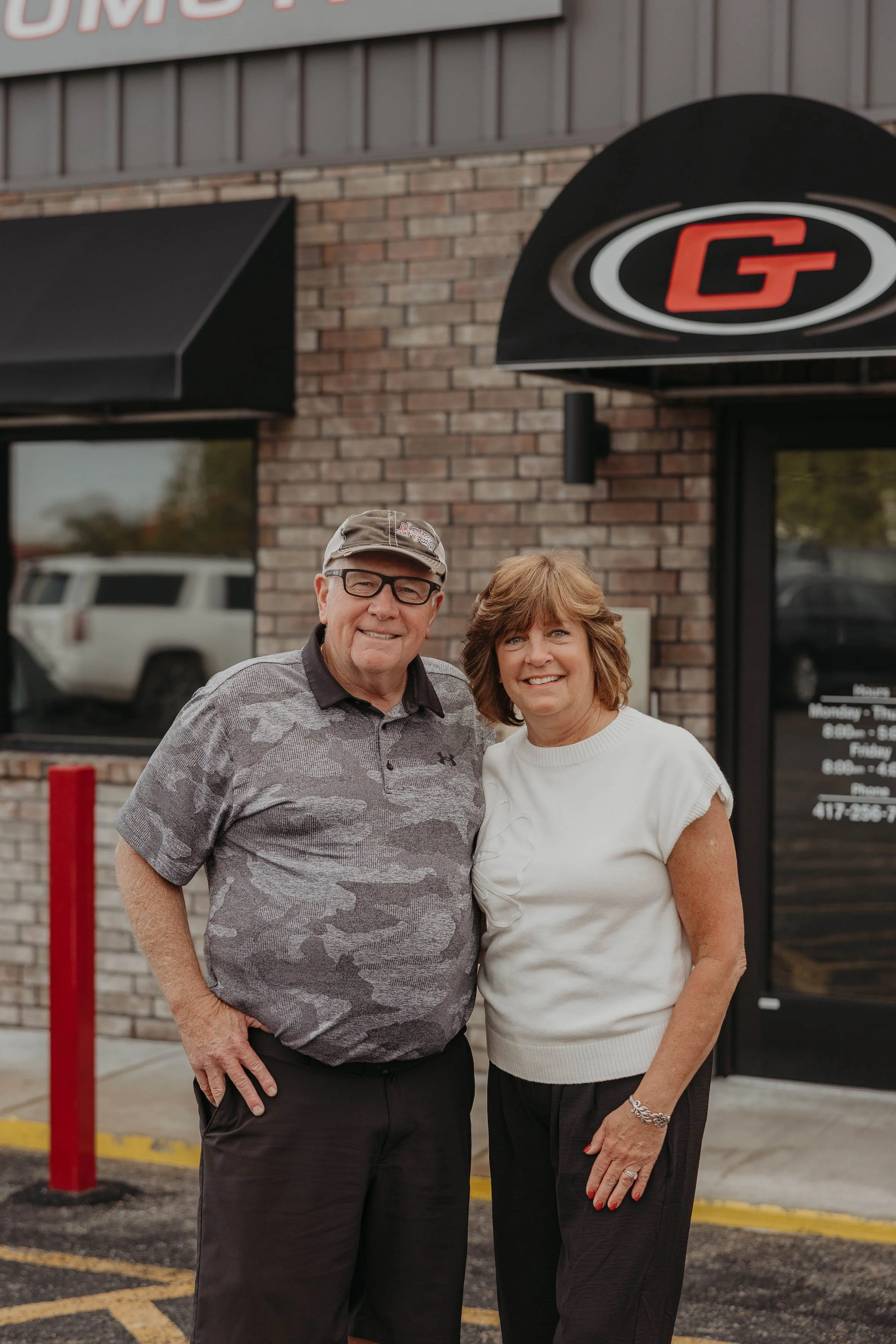 A smiling man and woman standing outside a brick building with a sign that has a red 'G' on a black background, in a parking lot.