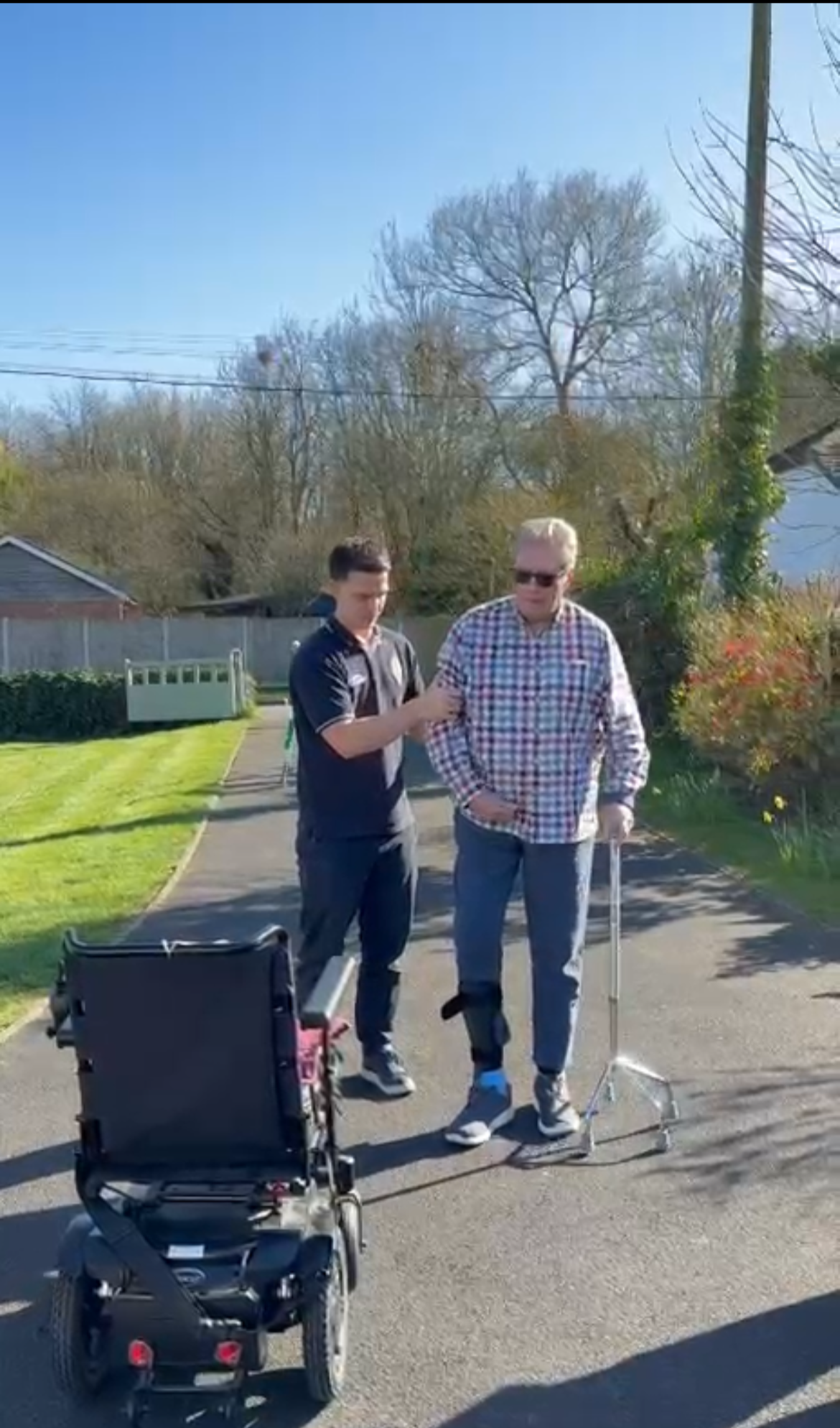 A young man stands next to an older man with a cane on a sidewalk in a residential area, with a wheelchair nearby. The young man appears to be assisting or talking to the older man on a sunny day.