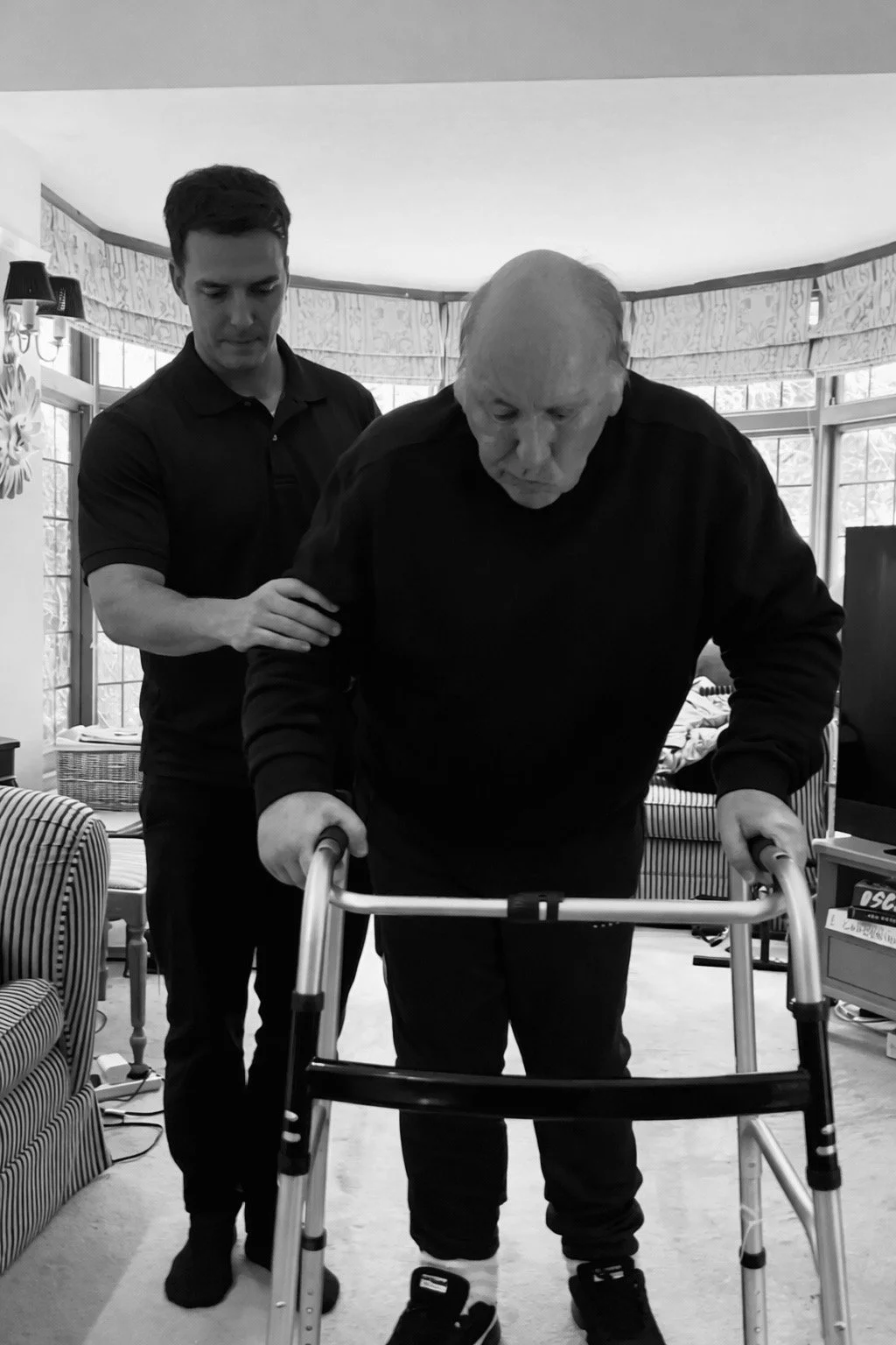An elderly man uses a walker with assistance from a young caregiver inside a living room with large windows and furniture.