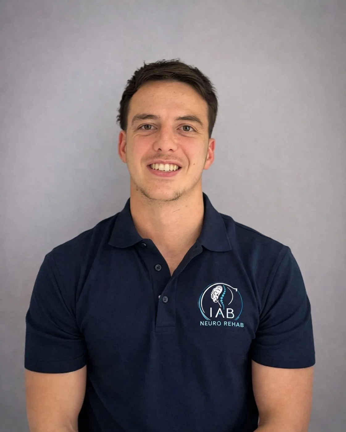 Young man smiling, wearing a navy polo shirt with a logo that reads 'IAB Neuro Rehab' and features a brain spine illustration, against a plain gray background.