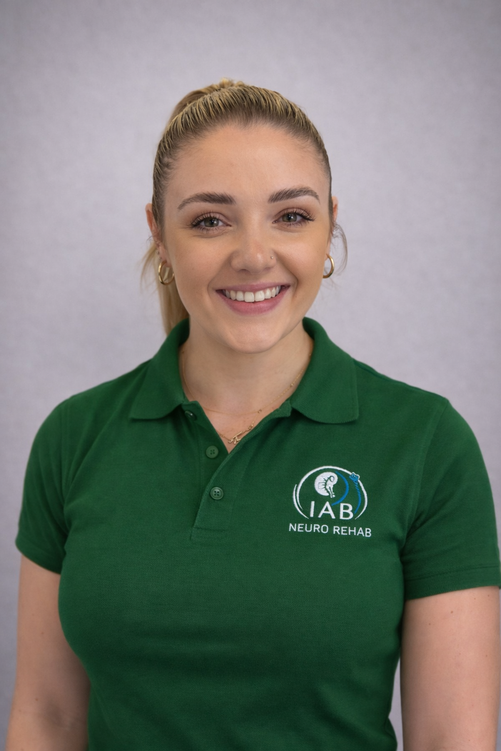 A young woman with blonde hair tied in a ponytail, wearing a green polo shirt with the logo 'IAB Neuro Rehab,' smiling at the camera, standing against a plain gray background.