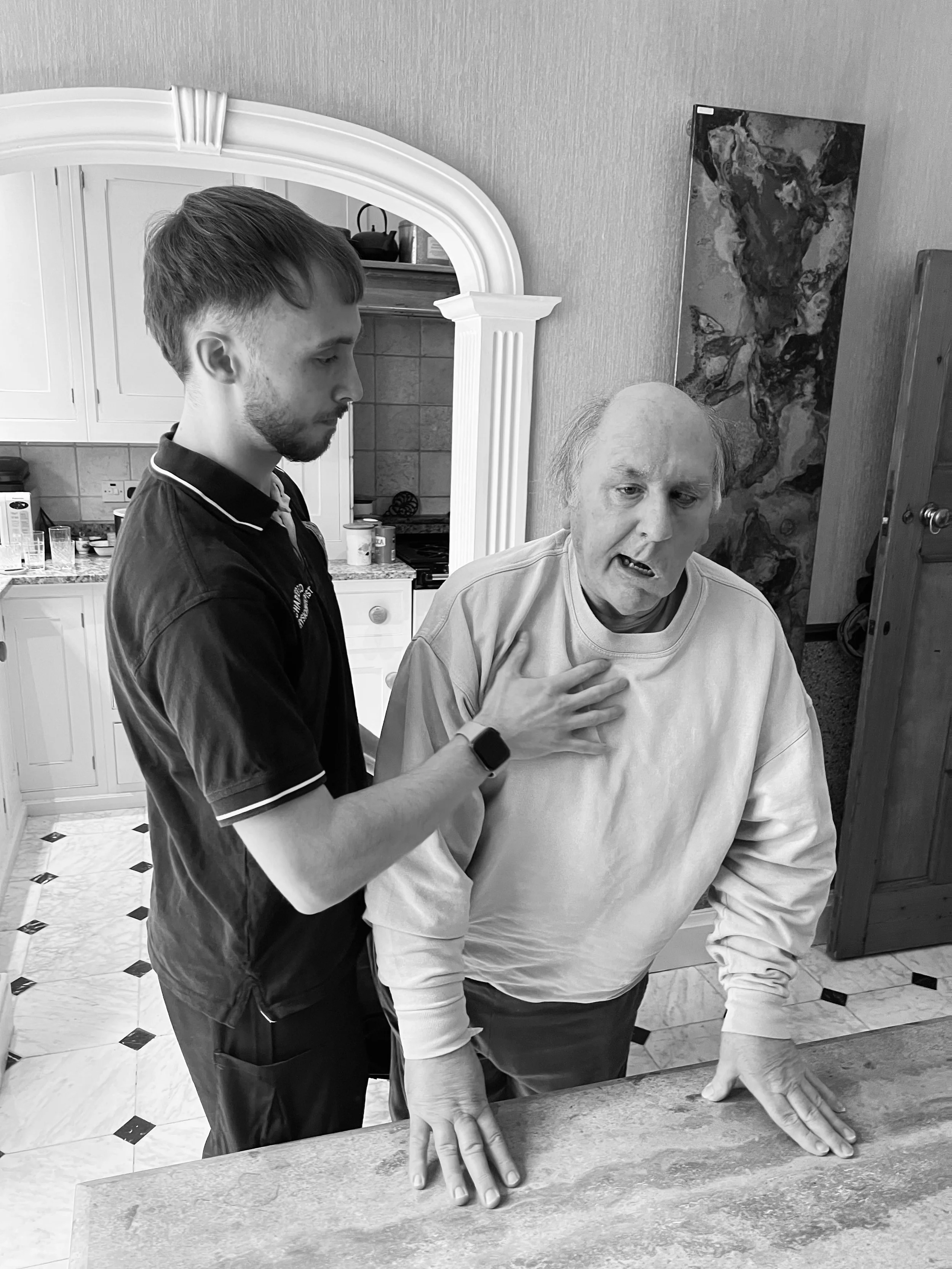 Young man with a smartwatch helping an older man who appears distressed and is holding his chest, in a kitchen.
