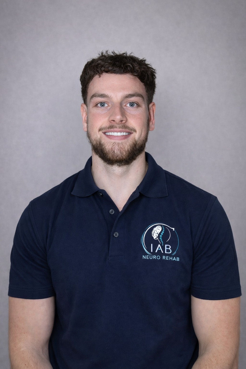 A young man with short dark curly hair, a beard, and blue eyes, smiling, wearing a navy blue polo shirt with a logo for IAB Neuro Rehab on the chest, standing against a plain grey background.
