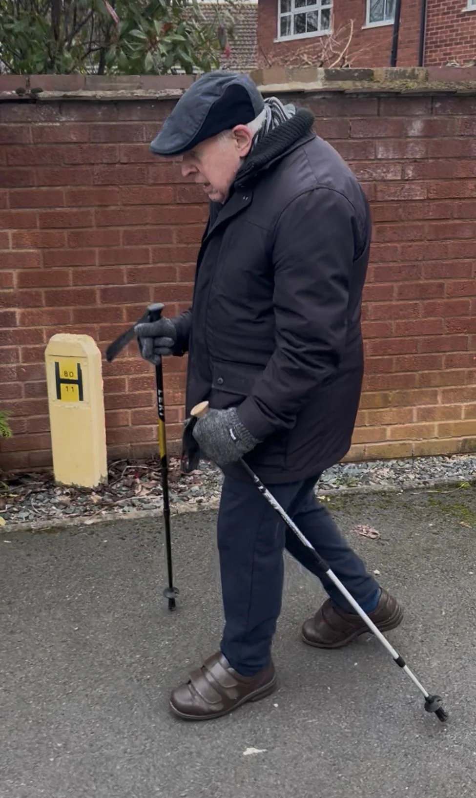 An elderly man walking outdoors on a paved path, using a walking stick, dressed in a black jacket, dark pants, brown shoes, gloves, and a cap, with a red brick wall and some shrubs in the background.