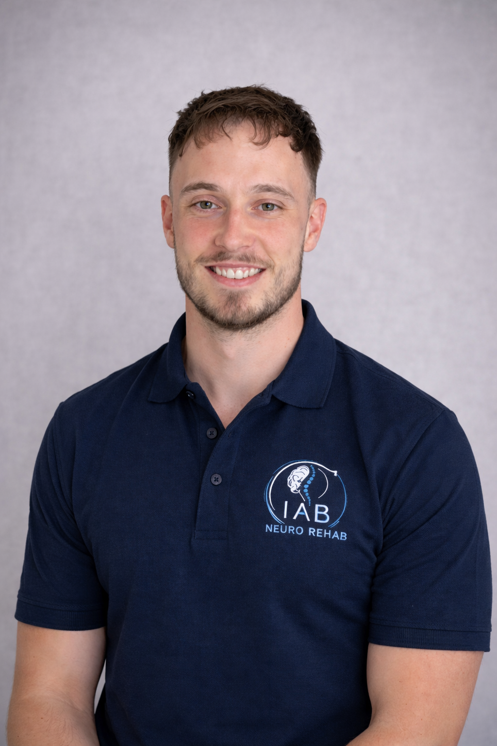 A smiling young man with short brown hair and a beard, wearing a navy blue polo shirt with an 'IAB Neuro Rehab' logo, posing against a plain light gray background.