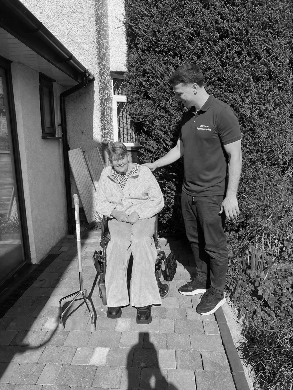 A young man in a polo shirt helping an elderly woman in a wheelchair outside near a house and bushes, smiling and interacting.