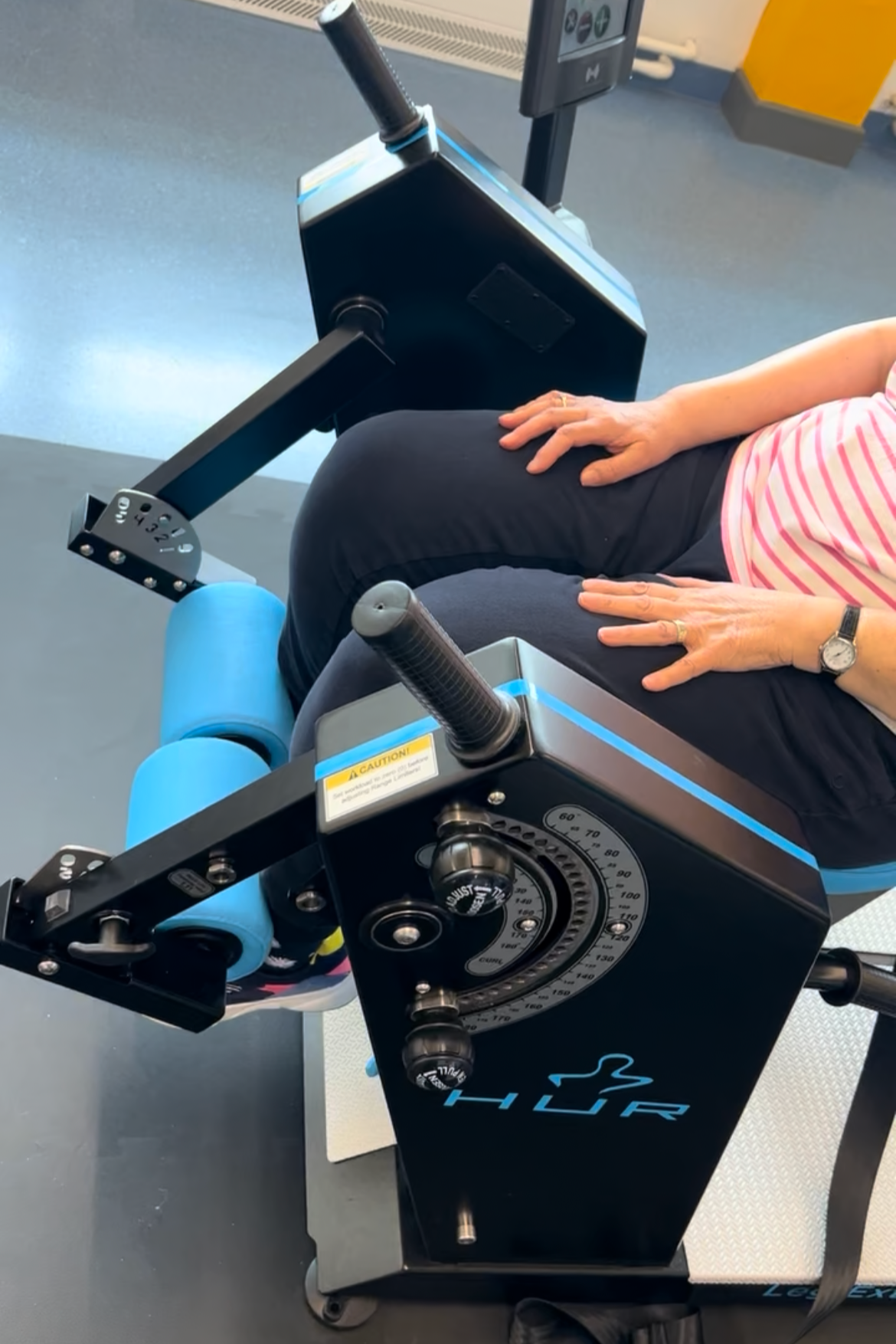 Close-up of a person using the HUR compression therapy device at a gym, with their hand on their thigh and the machine's control panel visible.