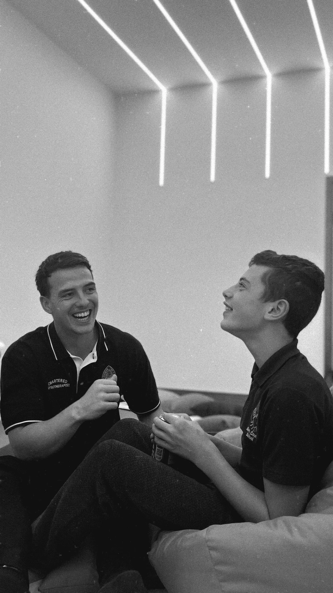 Two young men sitting on a couch, smiling and talking, with geometric neon lights on the ceiling in the background.