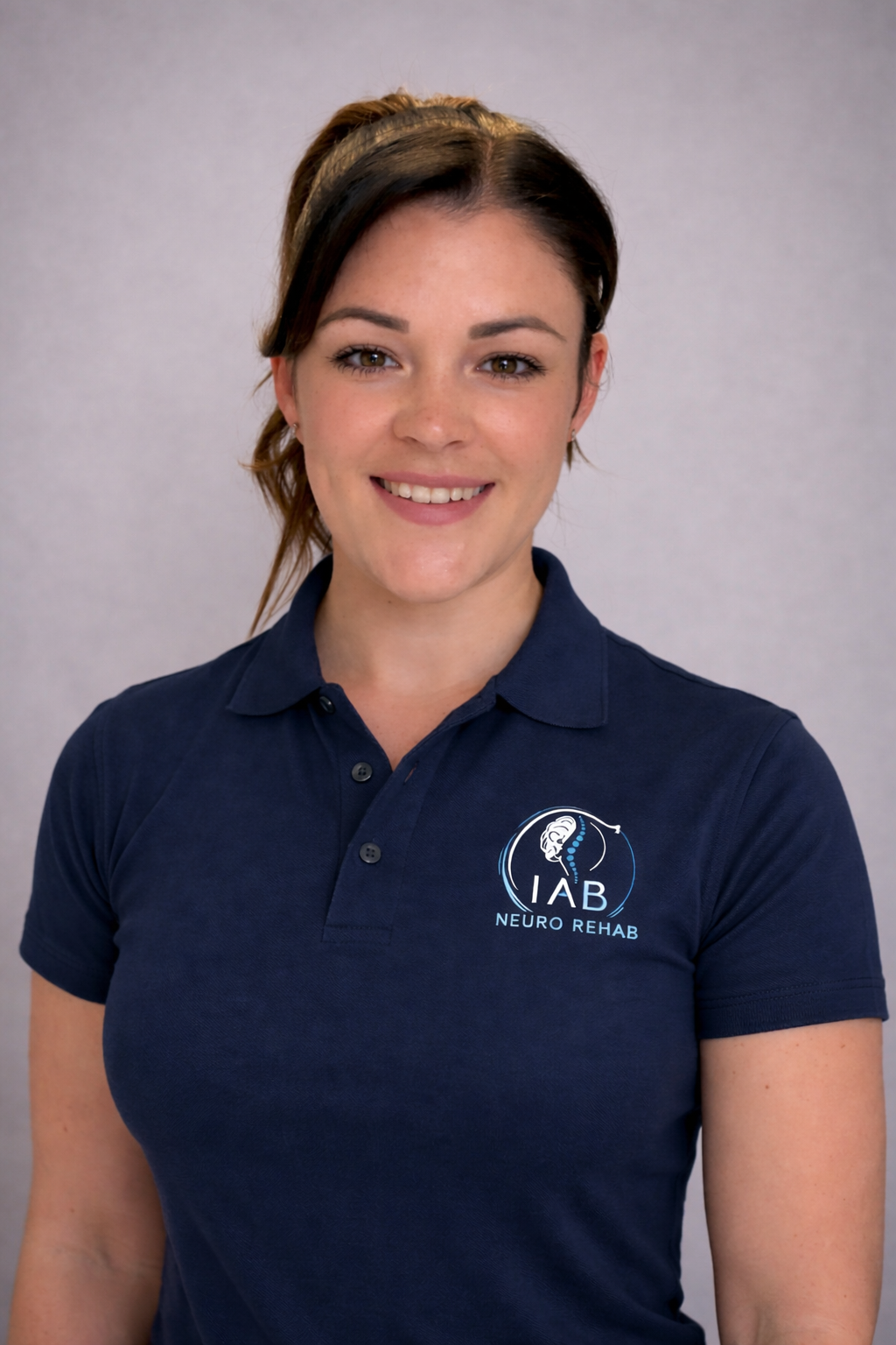 A young woman with brown hair tied back, wearing a navy blue polo shirt with a logo that reads "IAB Neuro Rehab," smiling at the camera against a light gray background.