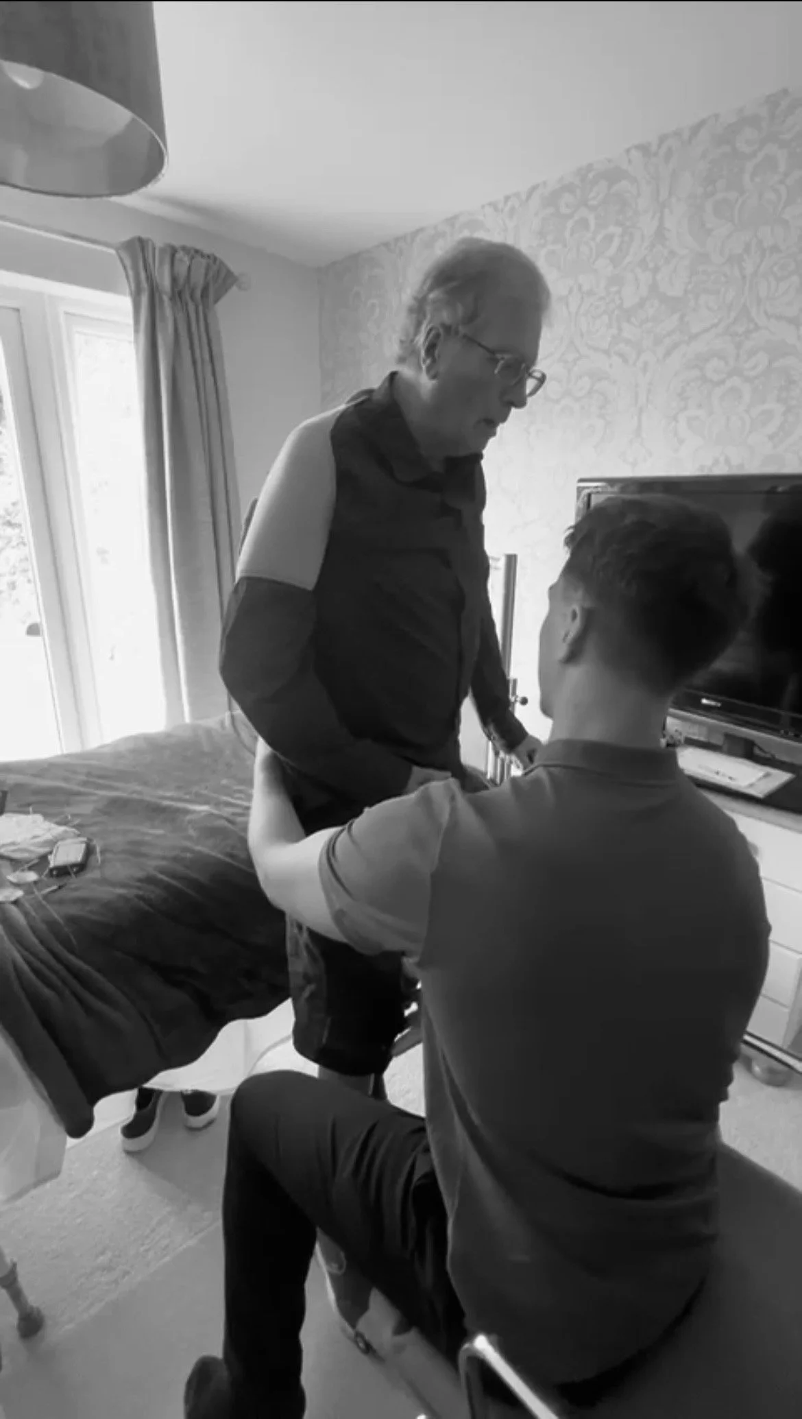 A black and white photo shows a nurse or doctor helping a young man into a hospital bed, with another older man observing. The setting appears to be a hospital room with a bed, television, and curtains.