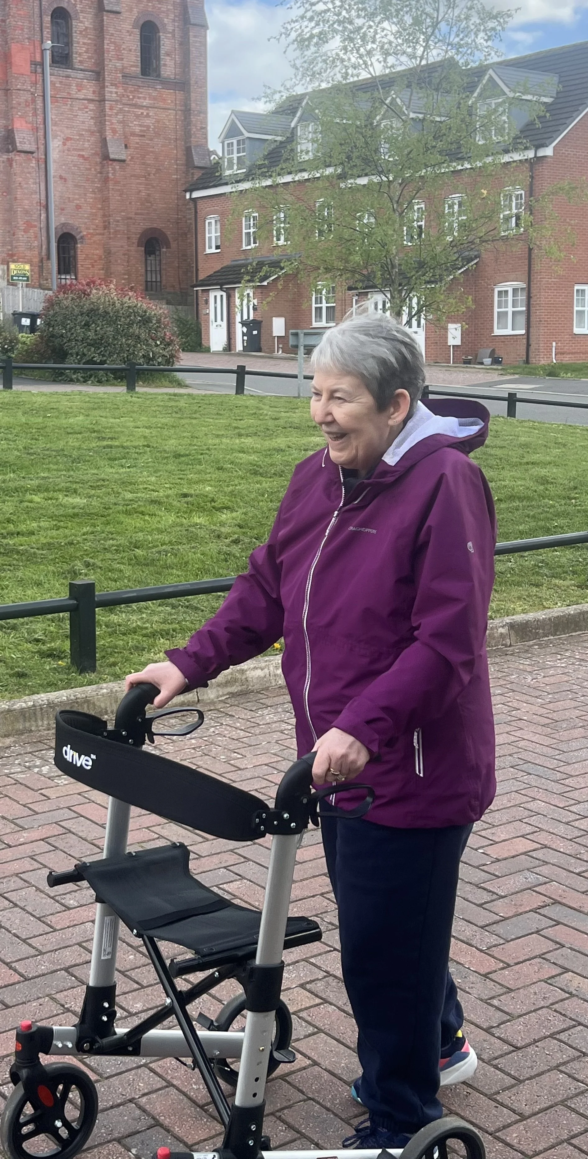 An elderly woman with short gray hair smiling while walking with a walker on a brick pathway in a park or residential area. She's wearing a purple jacket and dark pants, with trees, grass, and brick buildings in the background.