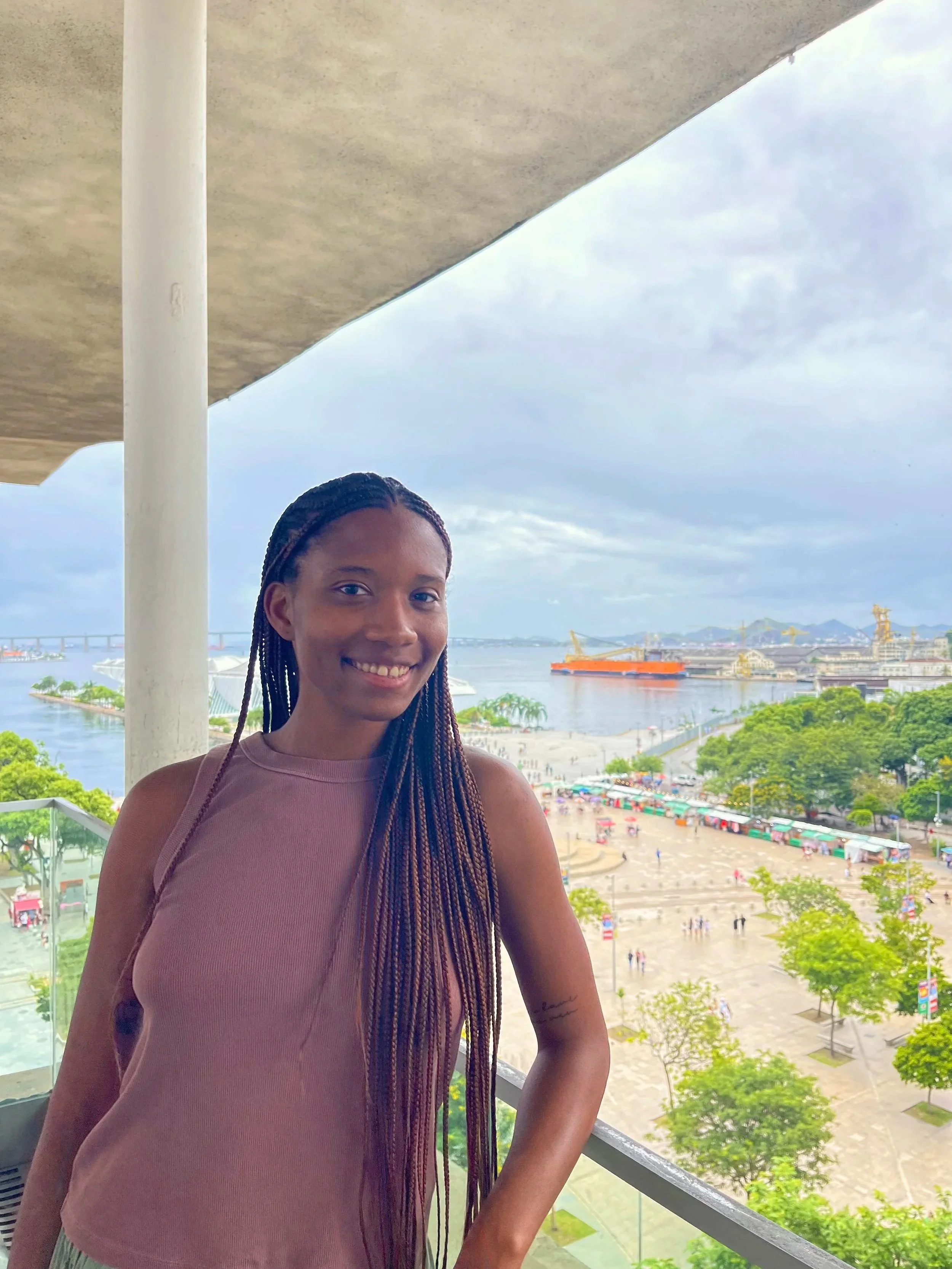 A woman with braided hair smiling on a balcony overlooking a cityscape with water, a bridge, and a beach with umbrellas.