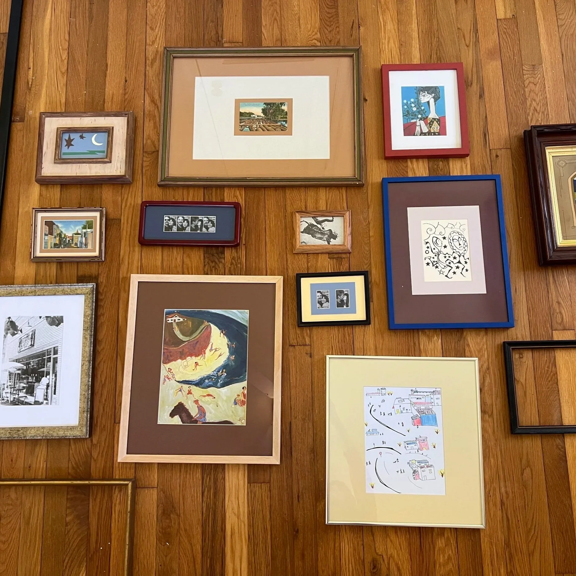 Various framed artworks, including abstract, landscape, and cartoon-style illustrations, hung on a wooden wall.