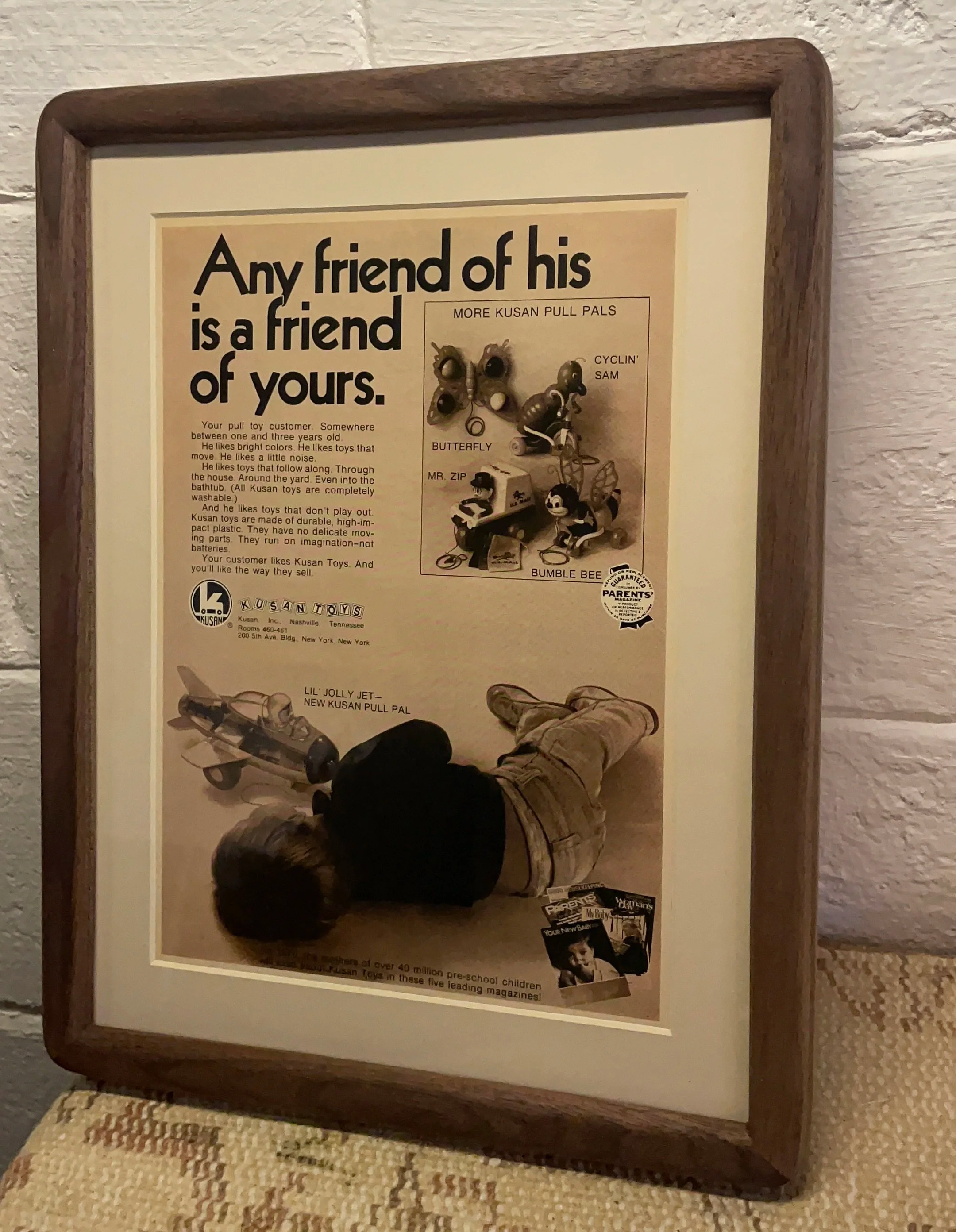Framed poster featuring a quote about Kusan pull toys, with a small photo of a child holding a toy, placed on a textured surface against a wall.