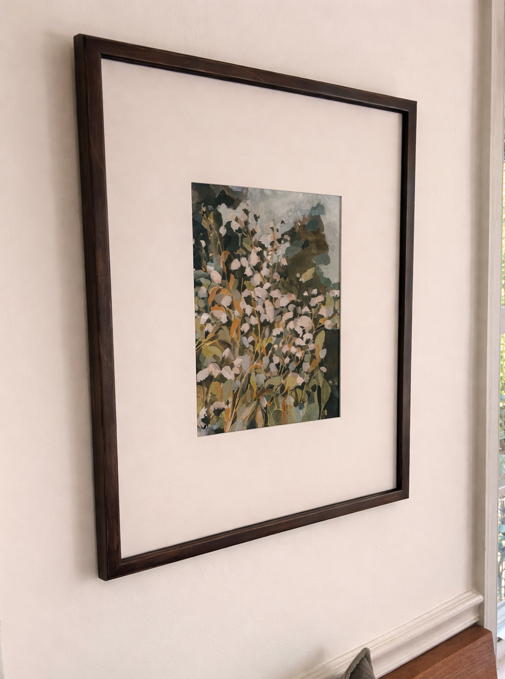 Framed floral painting with white and green blossoms on beige background hanging on a cream-colored wall near a window.