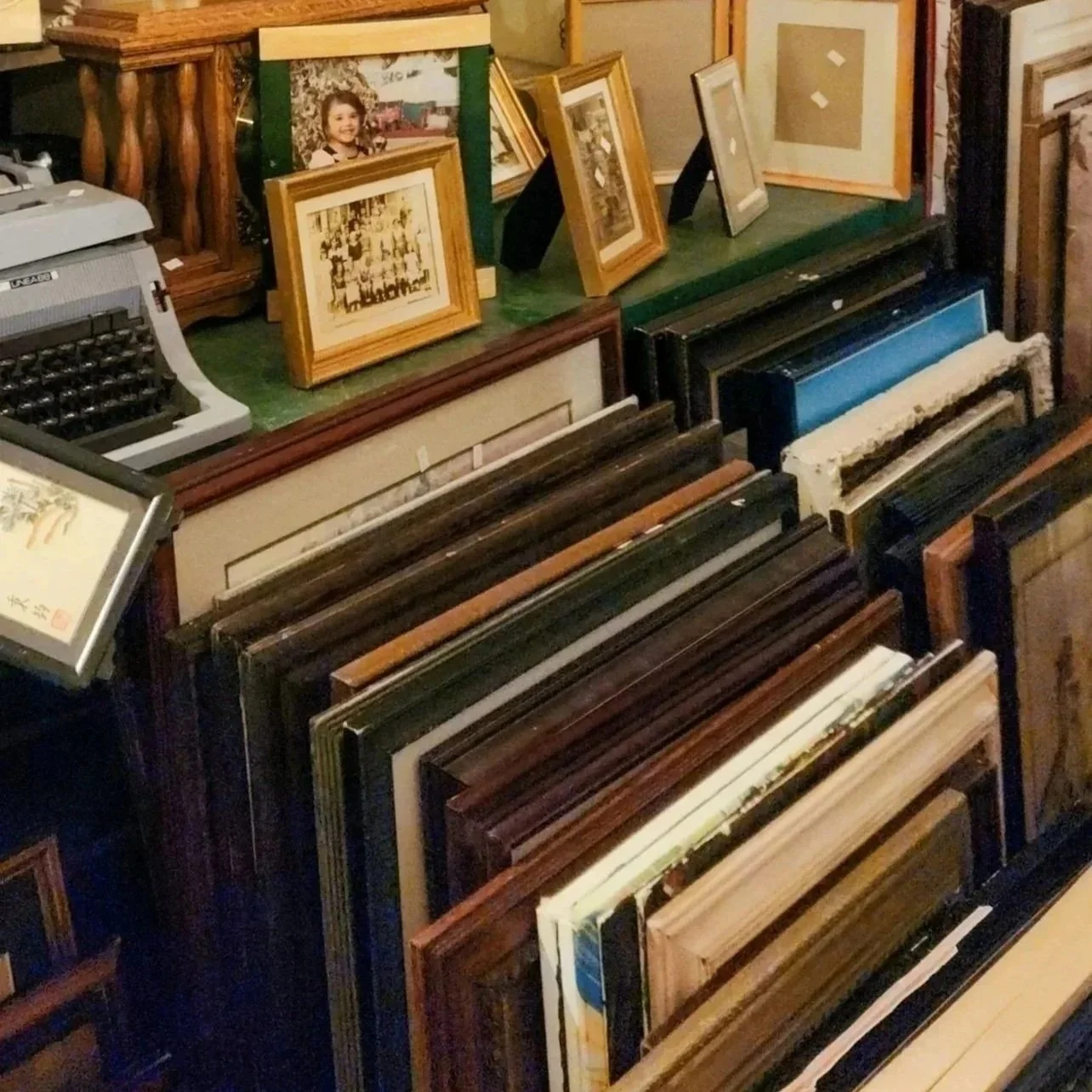 Various framed pictures and artwork leaning against a decorative wooden railing on a green surface, with a typewriter on the left and a large collection of framed artwork organized vertically.