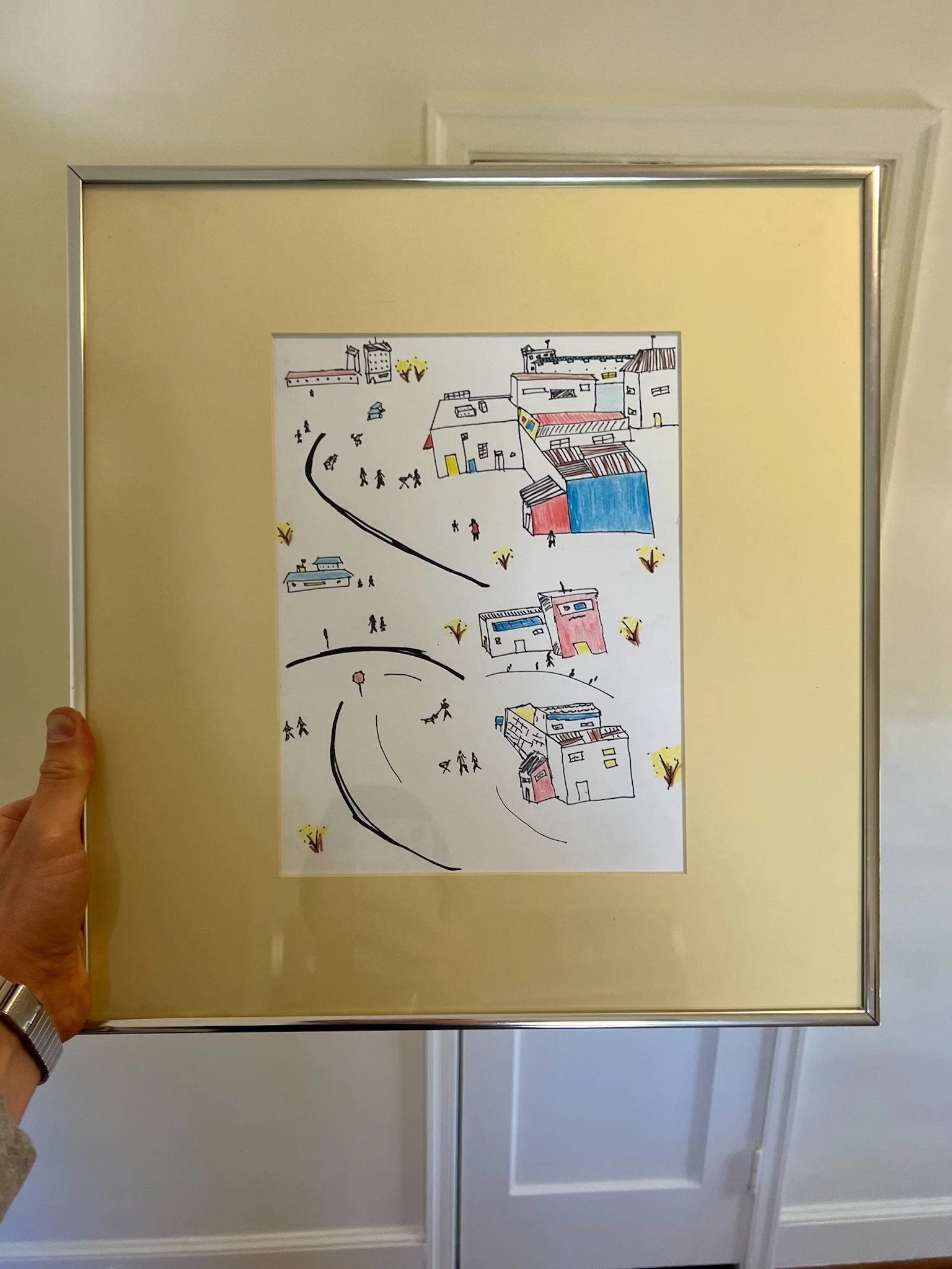 Framed drawing of an urban landscape with buildings, trees, and small figures, held in a hand against a door.