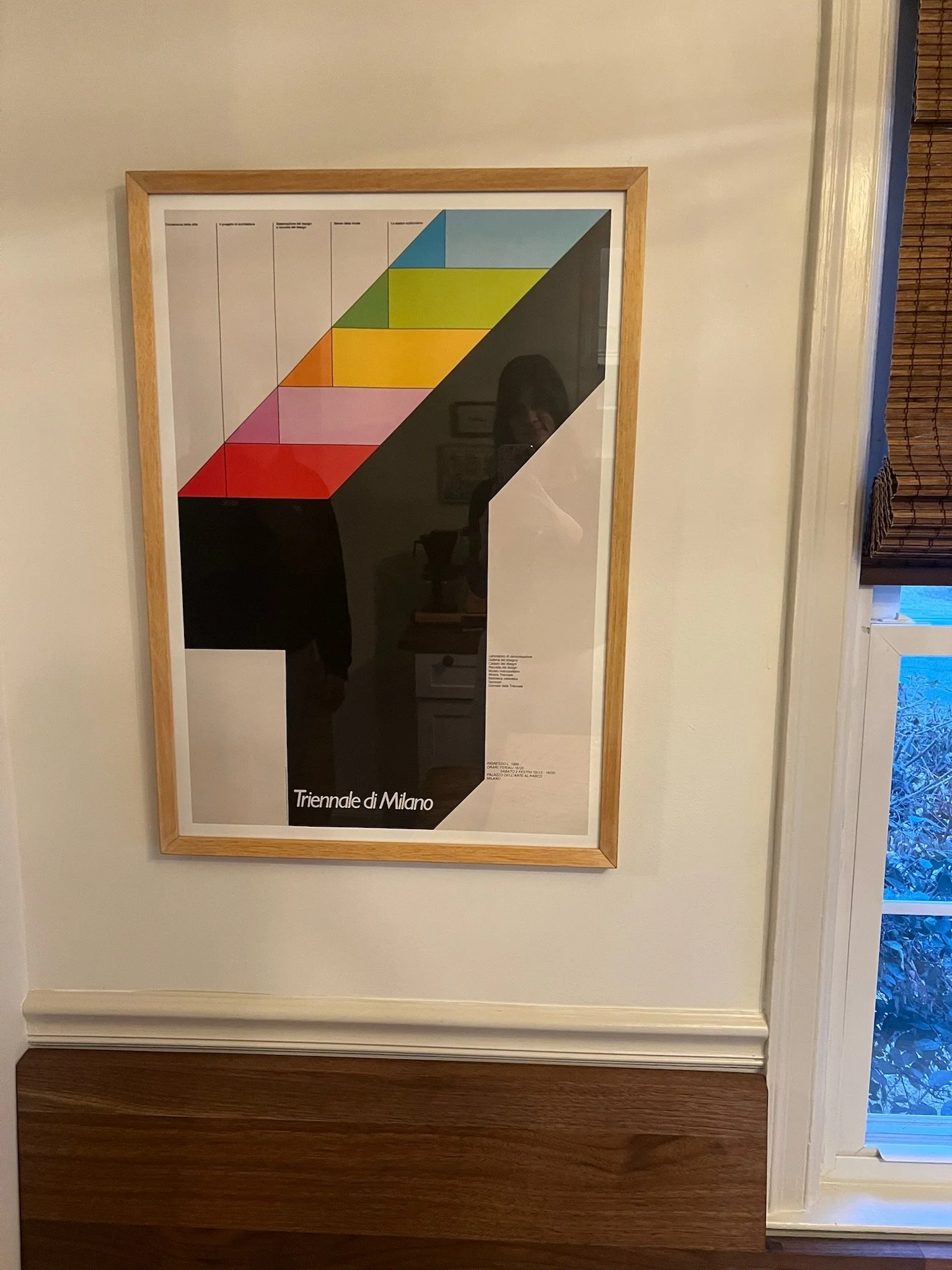 A framed poster on a white wall with a colorful geometric design and the text 'Triennale di Milano' at the bottom. A window with blinds is partially visible on the right side.