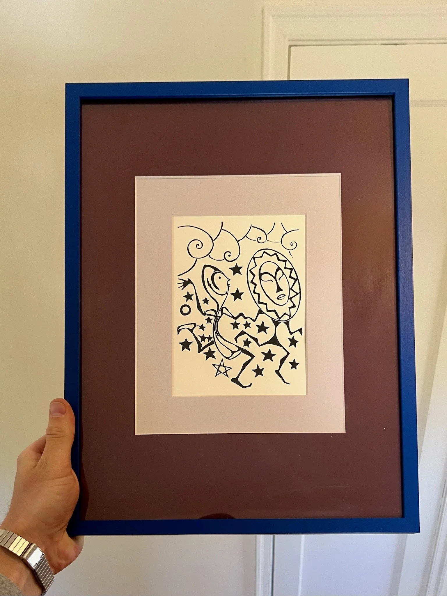 Hand holding a framed abstract black-and-white artwork with a cosmic theme, featuring a sun, moon, stars, and celestial faces, against a beige wall.