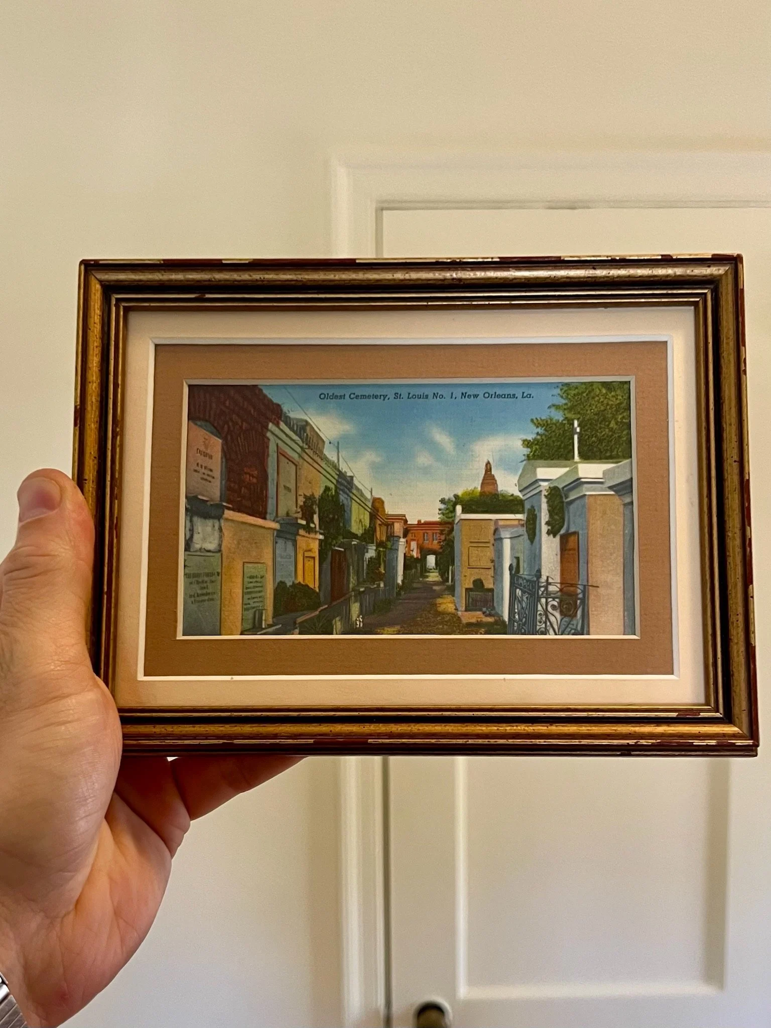 Framed painting of a cemetery scene with old tombstones, trees, and a brick building, with text indicating it's the oldest cemetery in New Orleans, Louisiana.