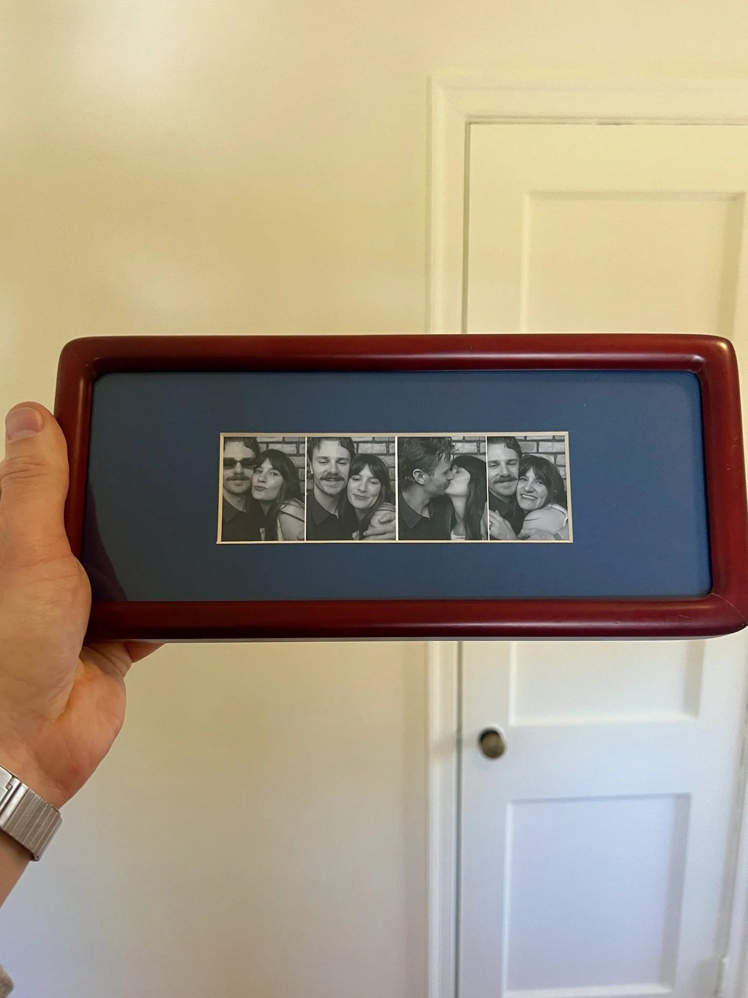 A person holding a small photo booth strip showing three pictures of a couple in black and white, with a white wall and door in the background.