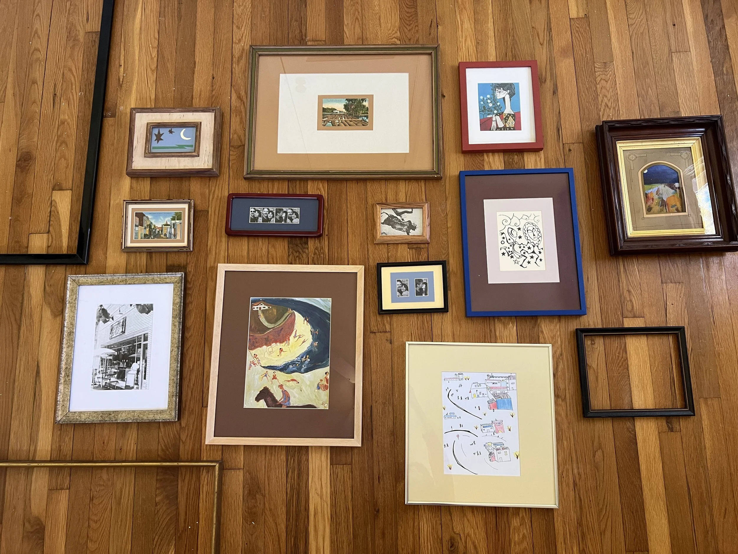 A gallery wall with various framed artworks on a wooden floor. The artworks include landscapes, portraits, abstract designs, and city scenes in different styles and frame colors.