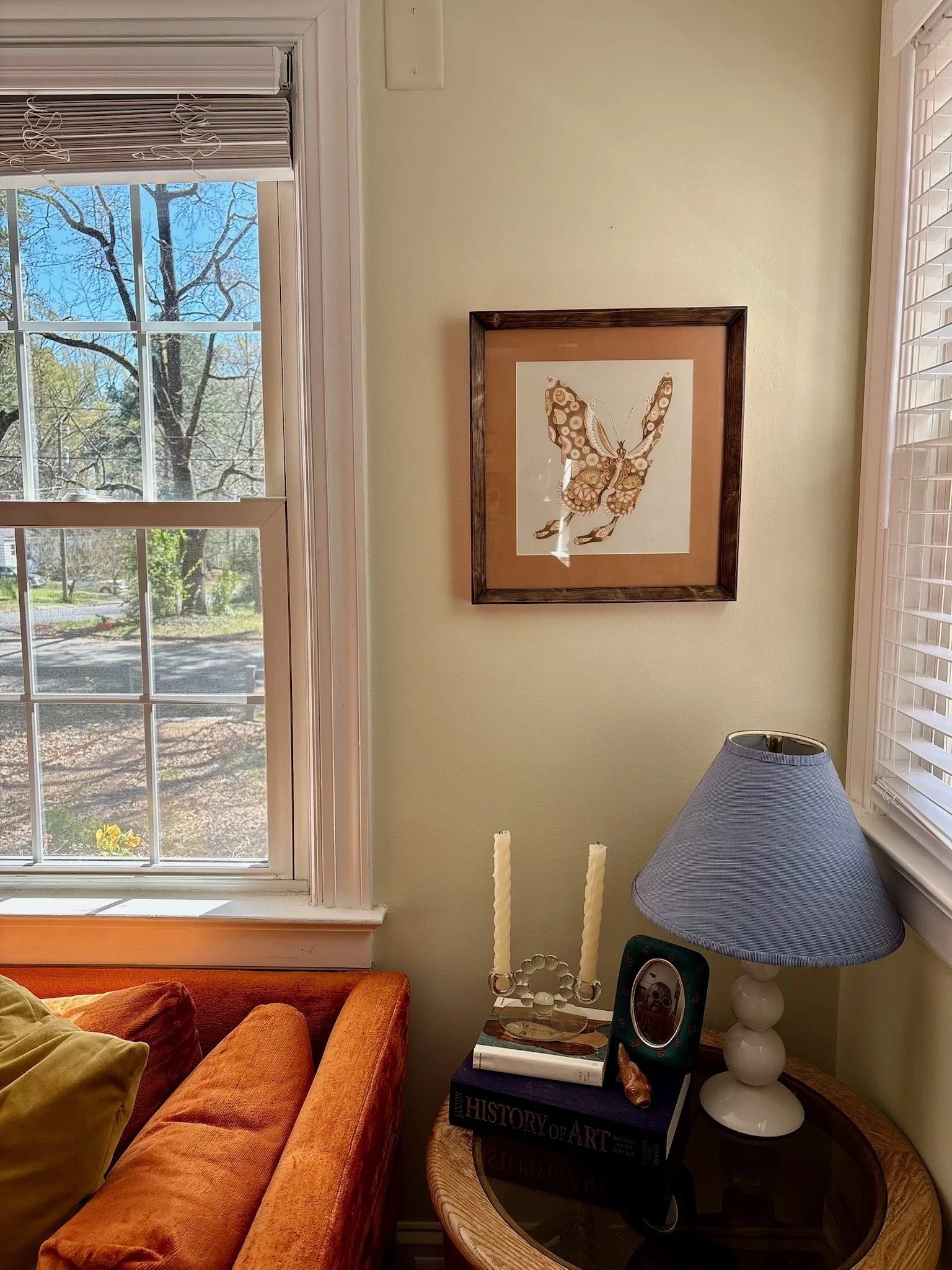 A cozy living room corner with a large window showing trees with budding leaves, a framed butterfly artwork on the wall, an orange sofa with a yellow pillow, a side table with candles, a black book titled "The History of Art," a small speaker, a lamp