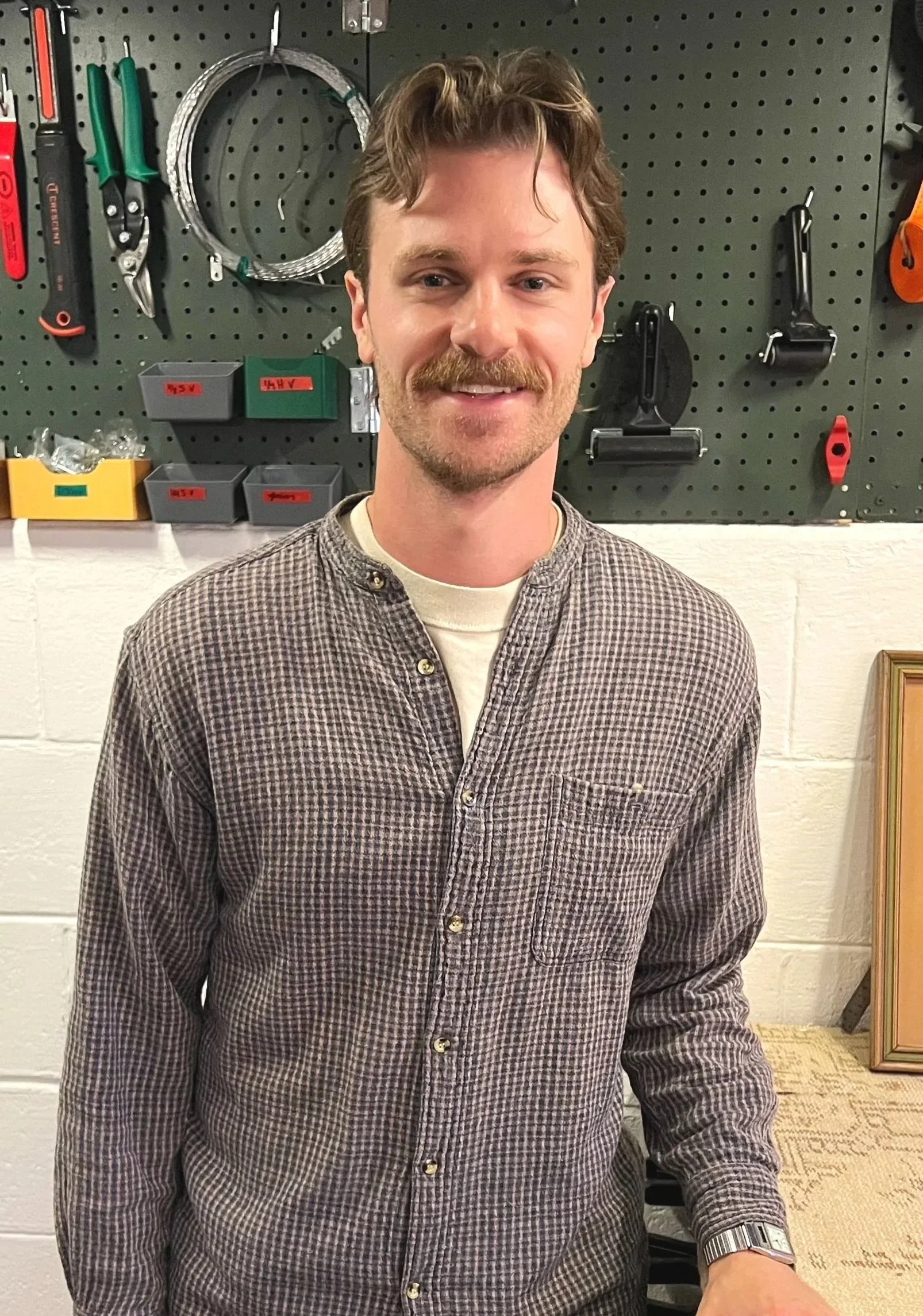 A man with light skin, brown wavy hair, and a mustache is smiling at the camera while standing in front of a black pegboard wall with various tools hanging on it. He's wearing a checkered shirt with a white undershirt and a wristwatch.