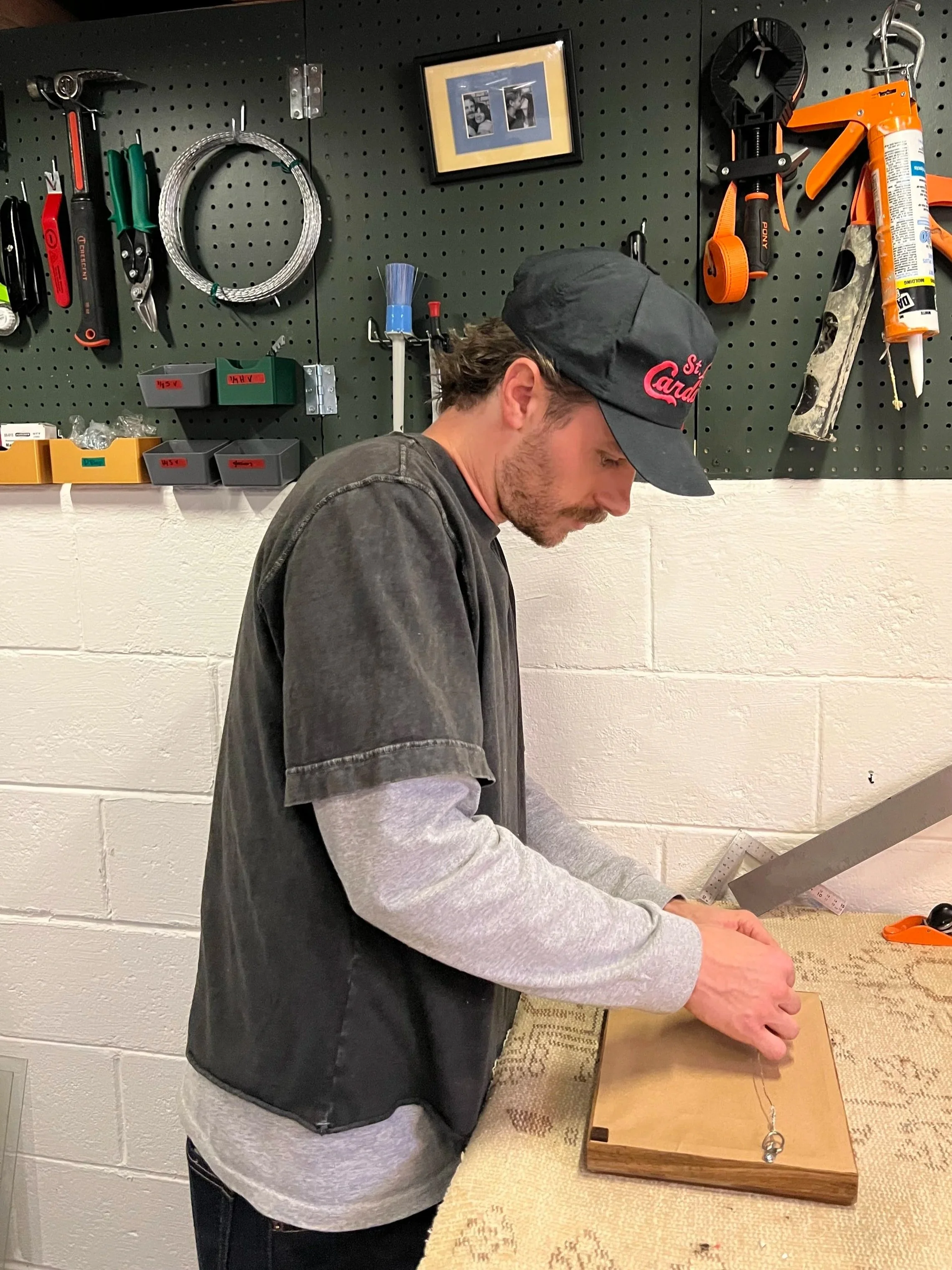 A man wearing a black cap and a gray sweatshirt working on a wooden box with a ruler inside a workshop. Behind him is a black pegboard with various tools hanging and a framed photo.