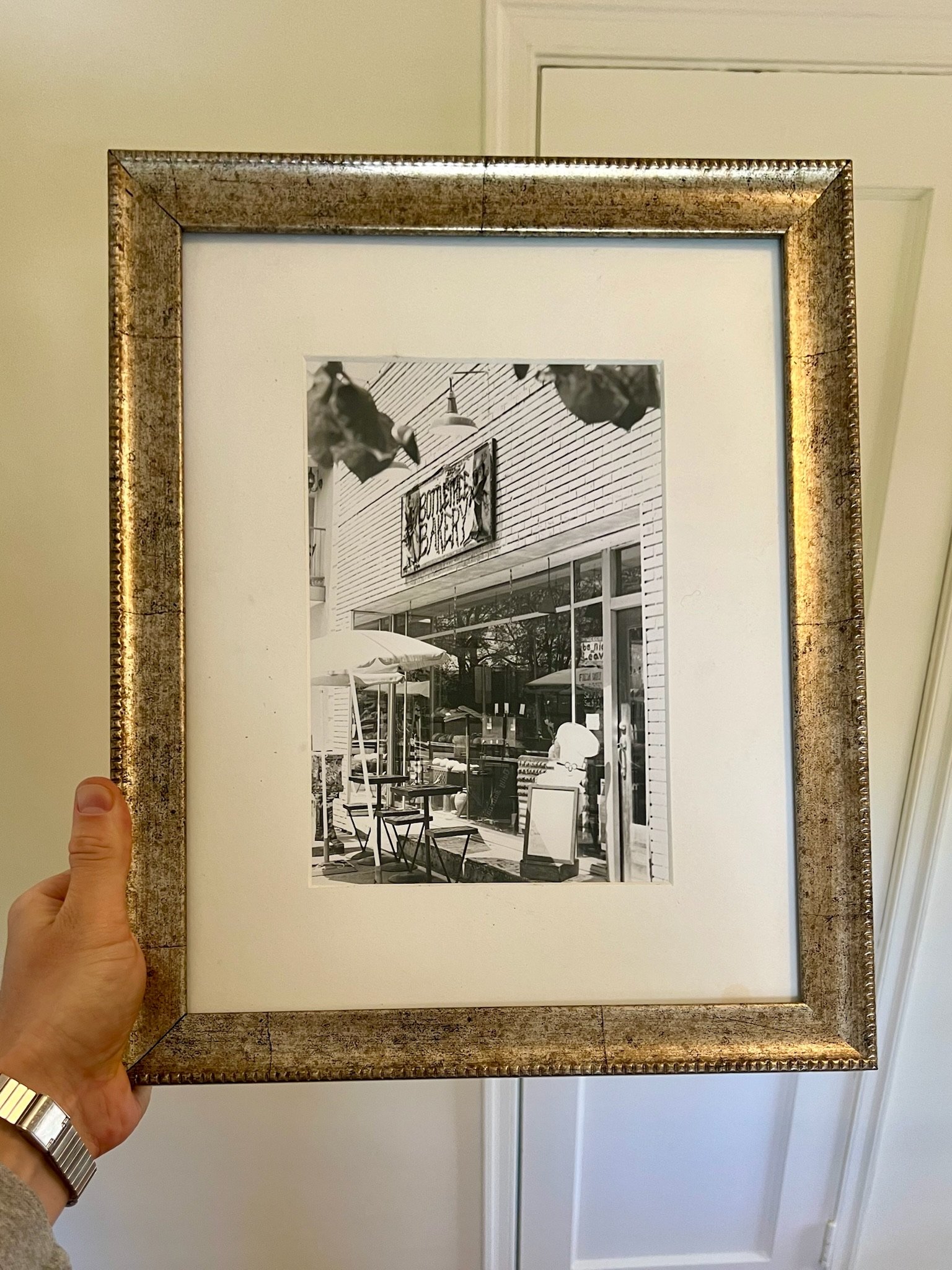 A person holding a framed black and white photograph of an outdoor cafe scene with tables, chairs, umbrellas, and a building with a sign that reads 'BOUTIQUE'.