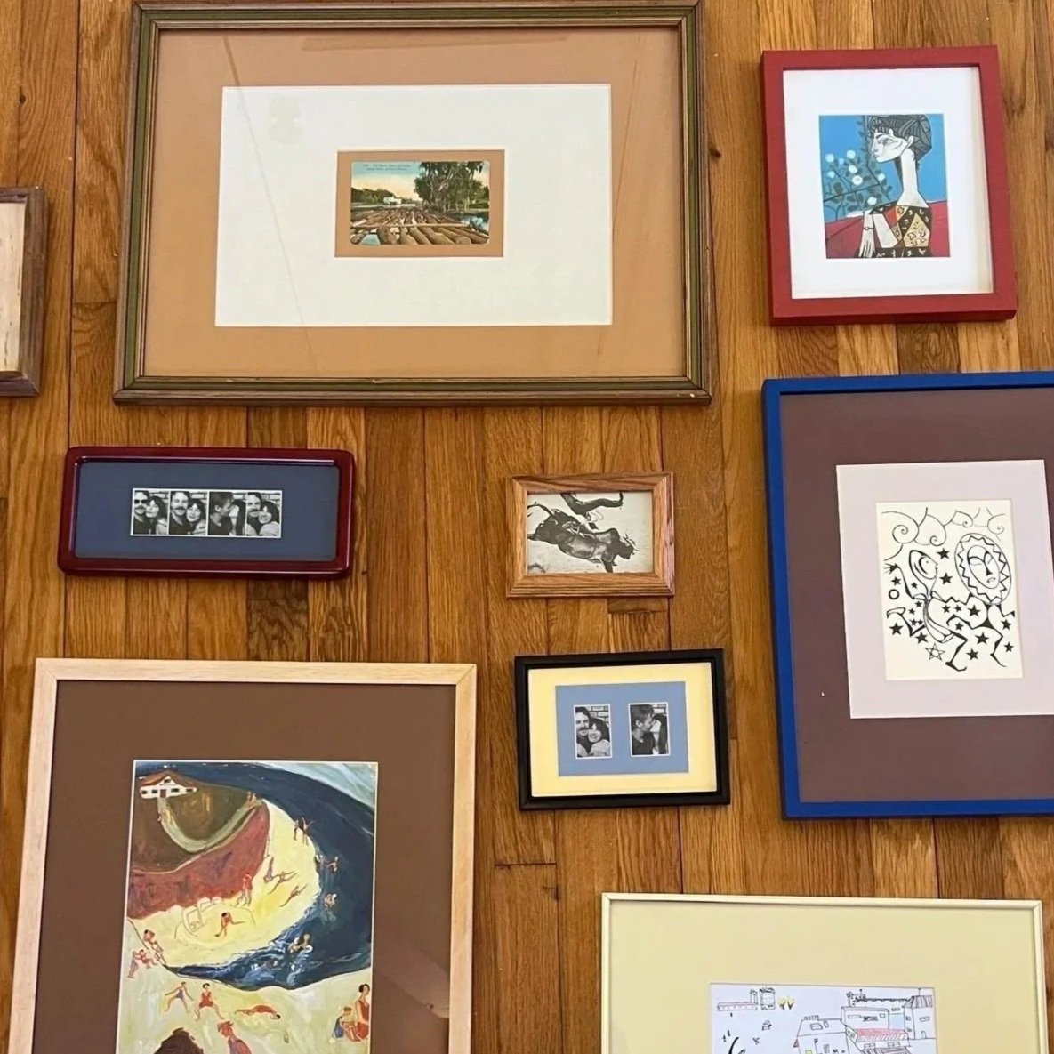 A collection of framed artwork and photographs arranged on a wooden wall, including paintings, sketches, and photo strips, in various sizes and frame colors.