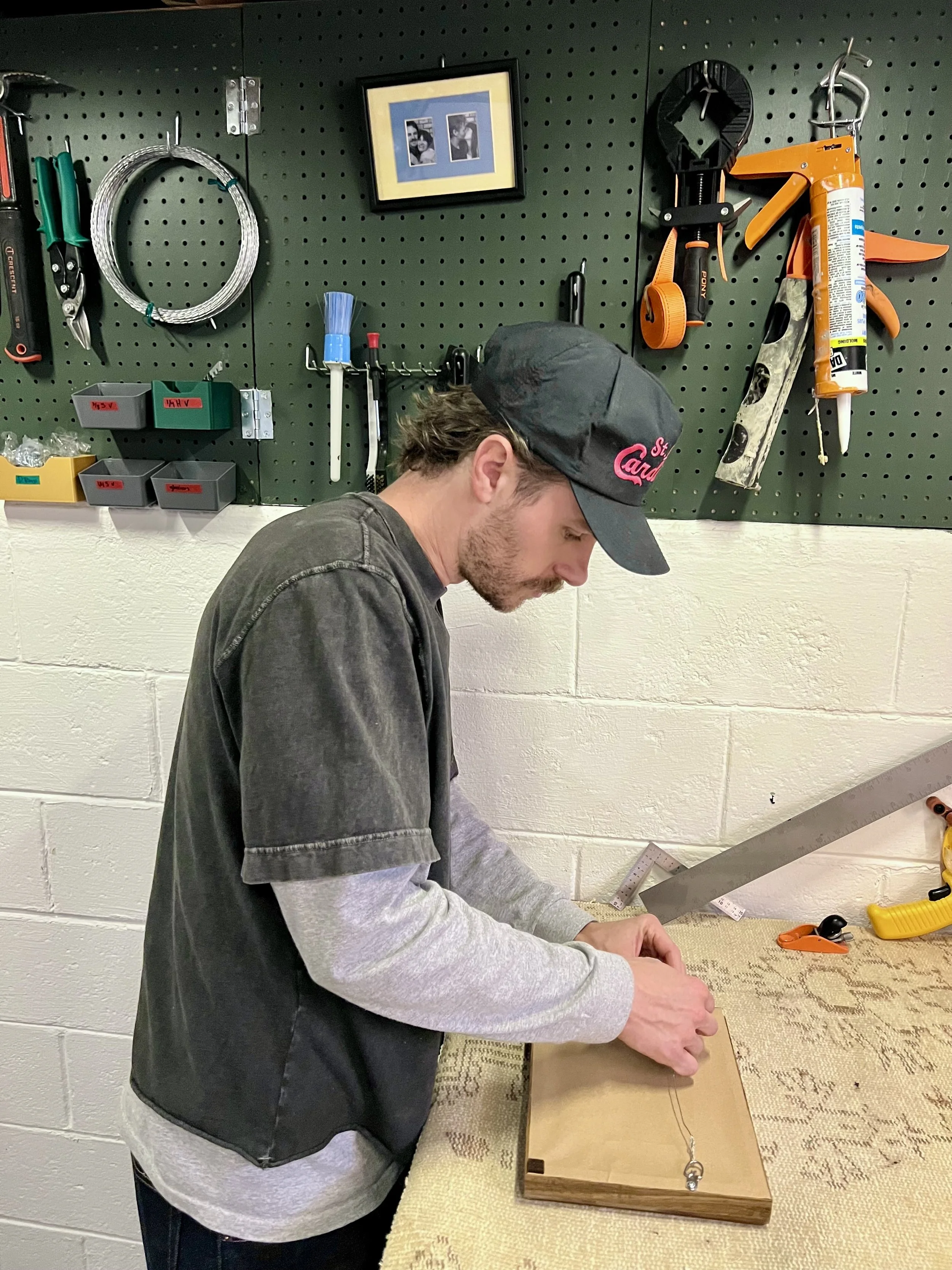 A young man wearing a black baseball cap and a gray long-sleeve shirt working on a woodworking project at a workbench in a garage workshop. Behind him is a pegboard with tools and accessories hanging.