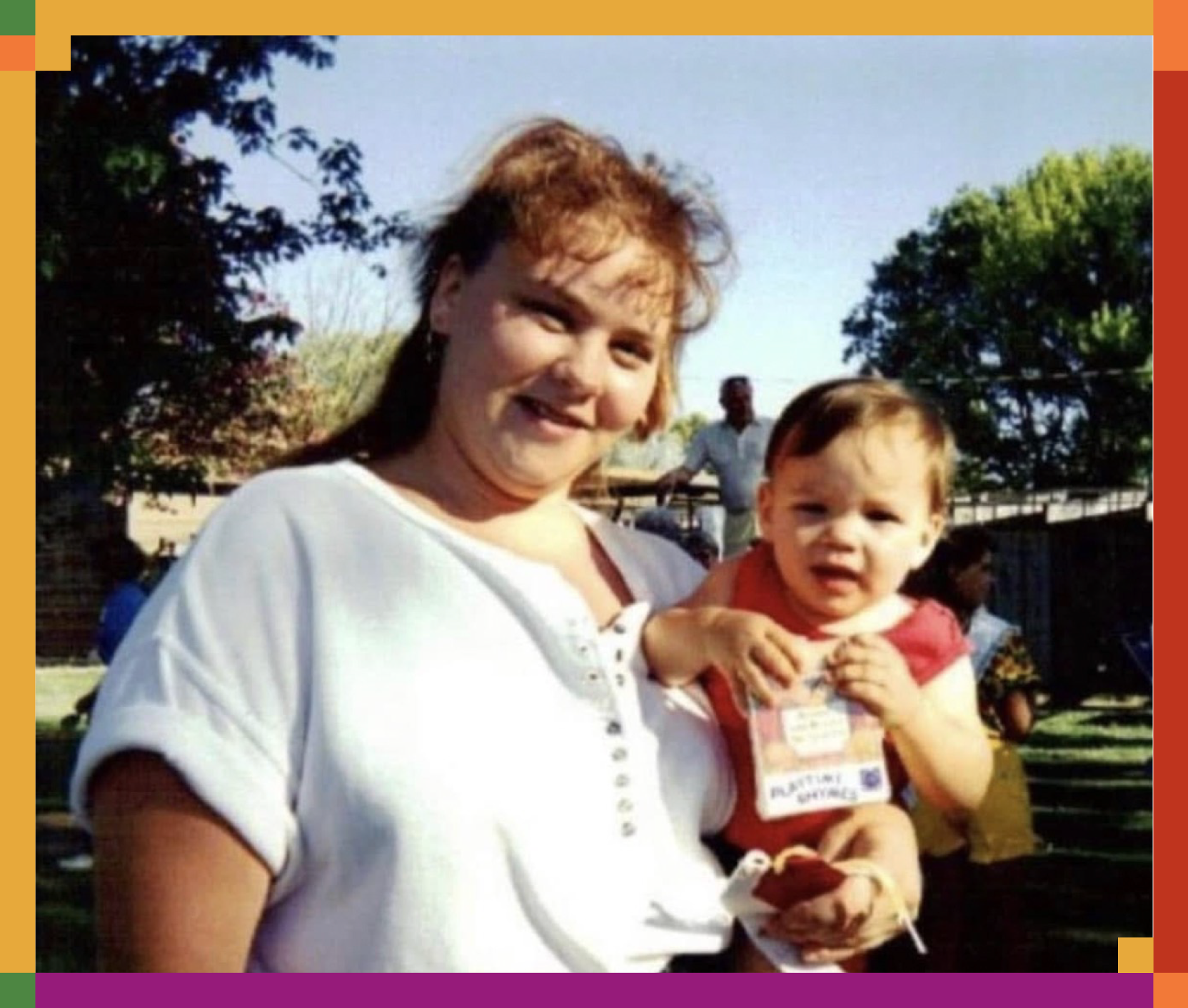 A woman holding a young child outdoors in a park or garden area, with trees and a person in the background.