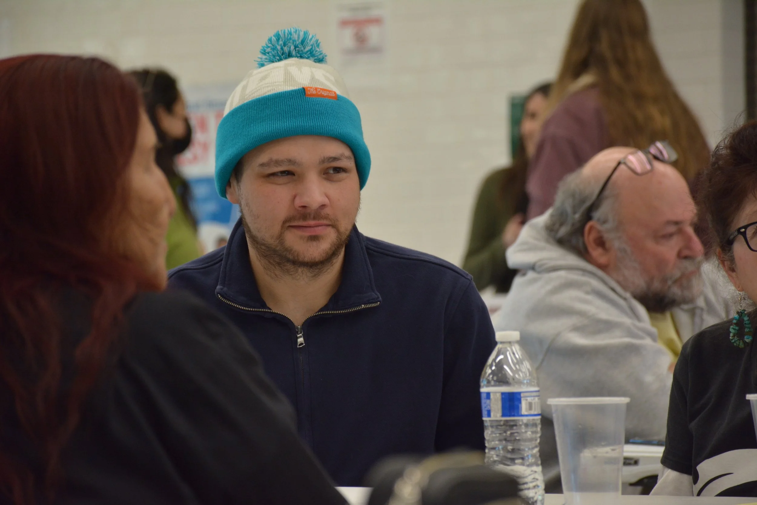 A young man wearing a multicolored beanie hat and a navy zip-up jacket, sitting at a table in a crowded indoor setting.