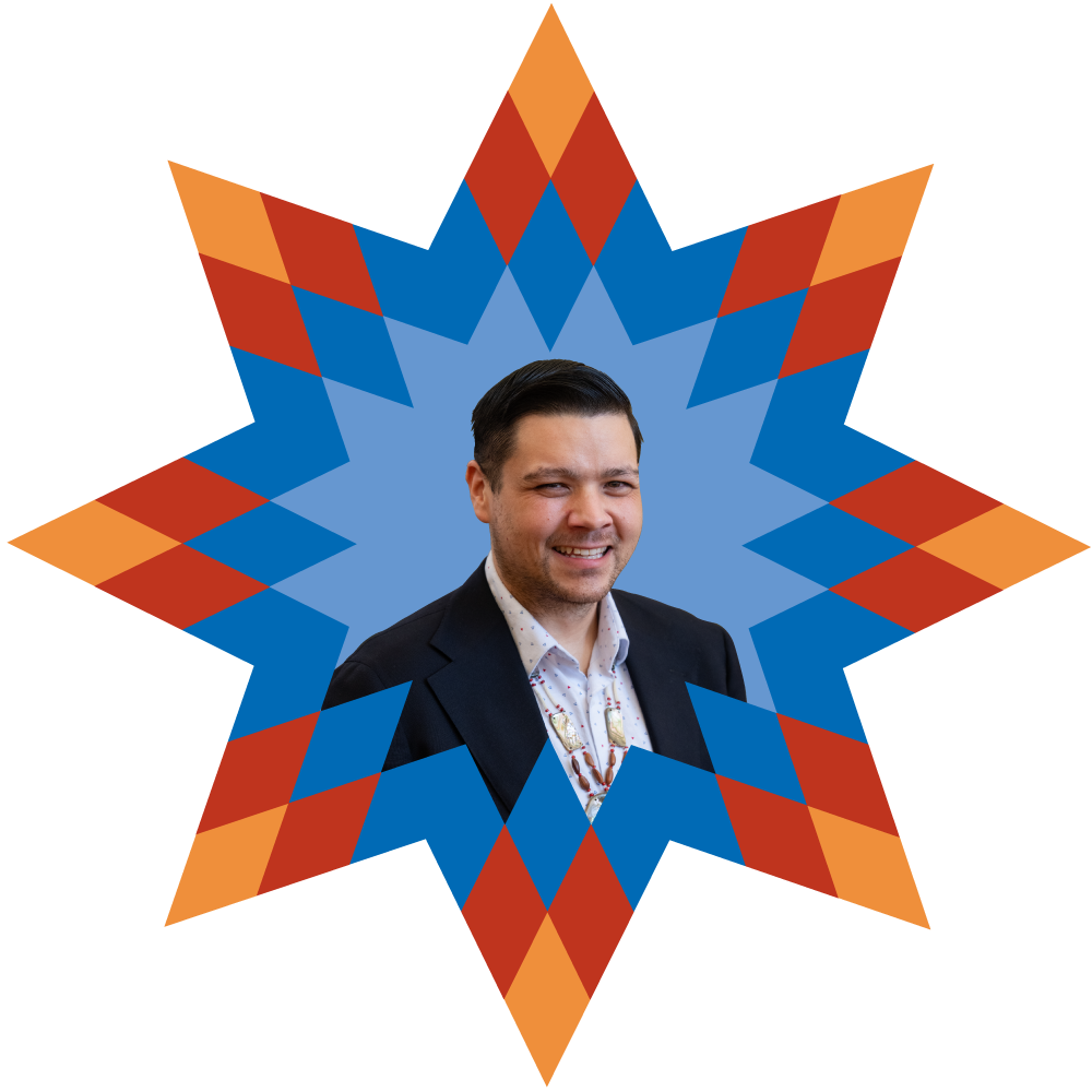 A man in a suit smiling and winking, with a colorful, geometric background resembling a star or explosion pattern.