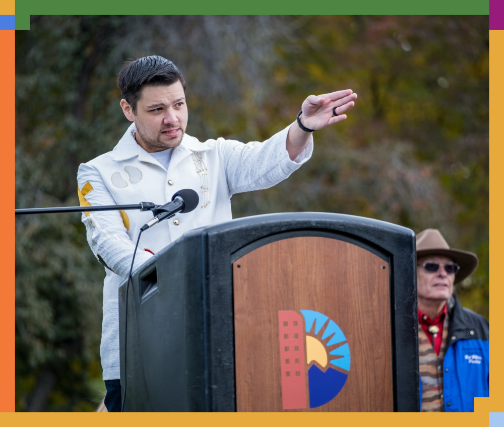 A man speaking at a podium outdoors, gesturing with his right hand while another man observes in the background.