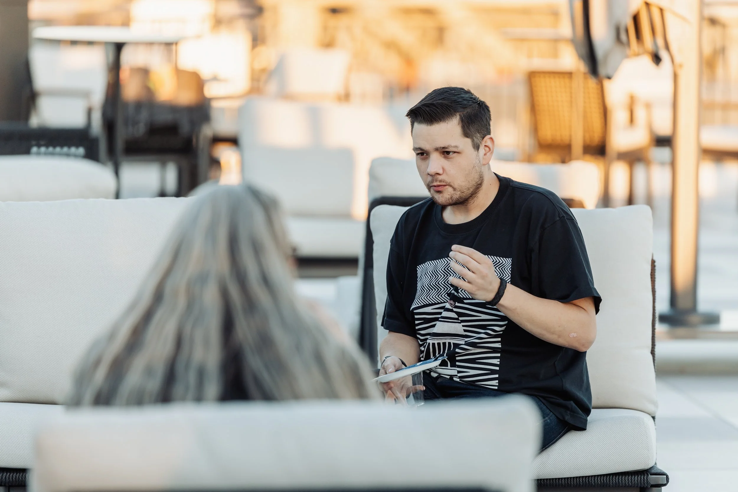 A young man with short dark hair and a beard sitting on a white outdoor sofa, holding a tablet and a glass, engaged in conversation with a woman with long gray hair, outdoors during daylight.
