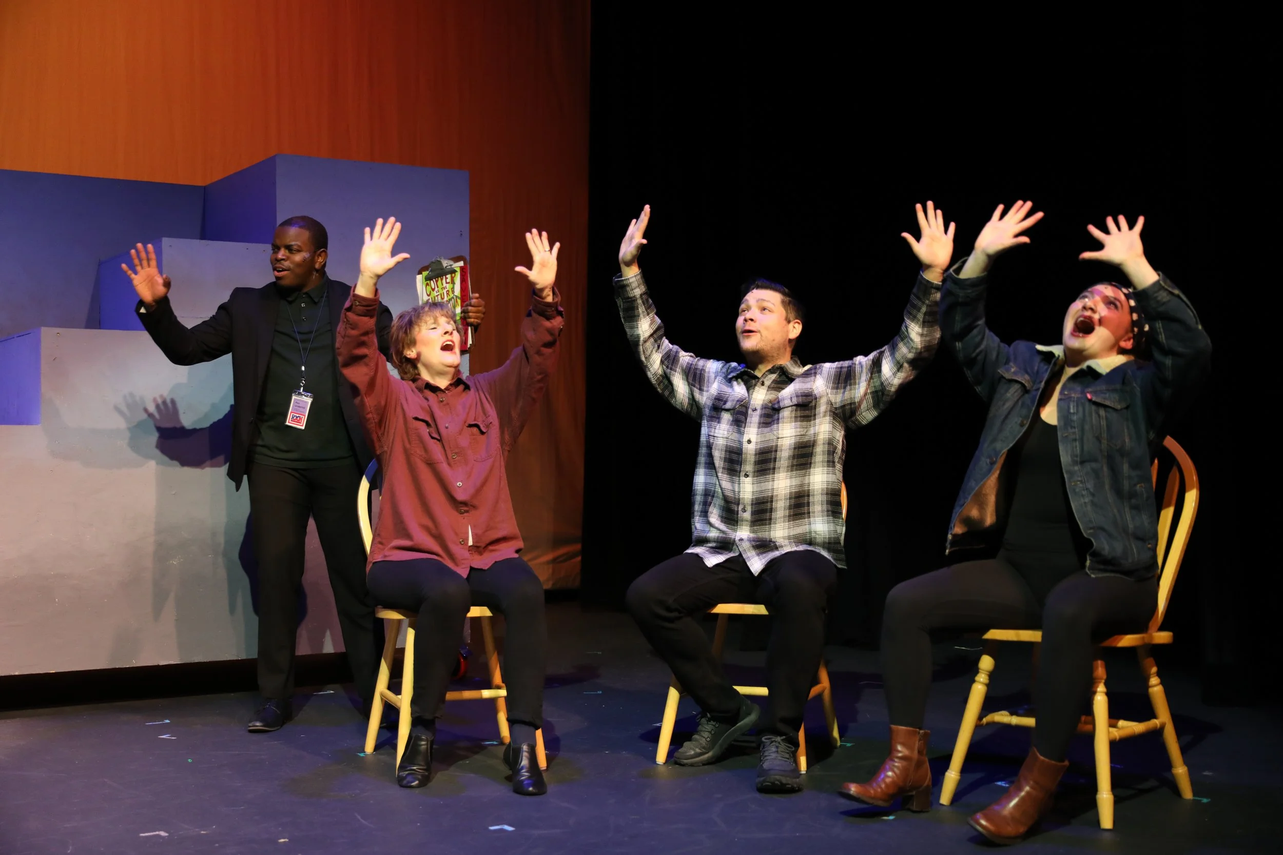 Four actors performing on a stage, three seated in chairs with their arms raised, with a man standing behind them holding a script. The scene appears to be part of a theatrical play.