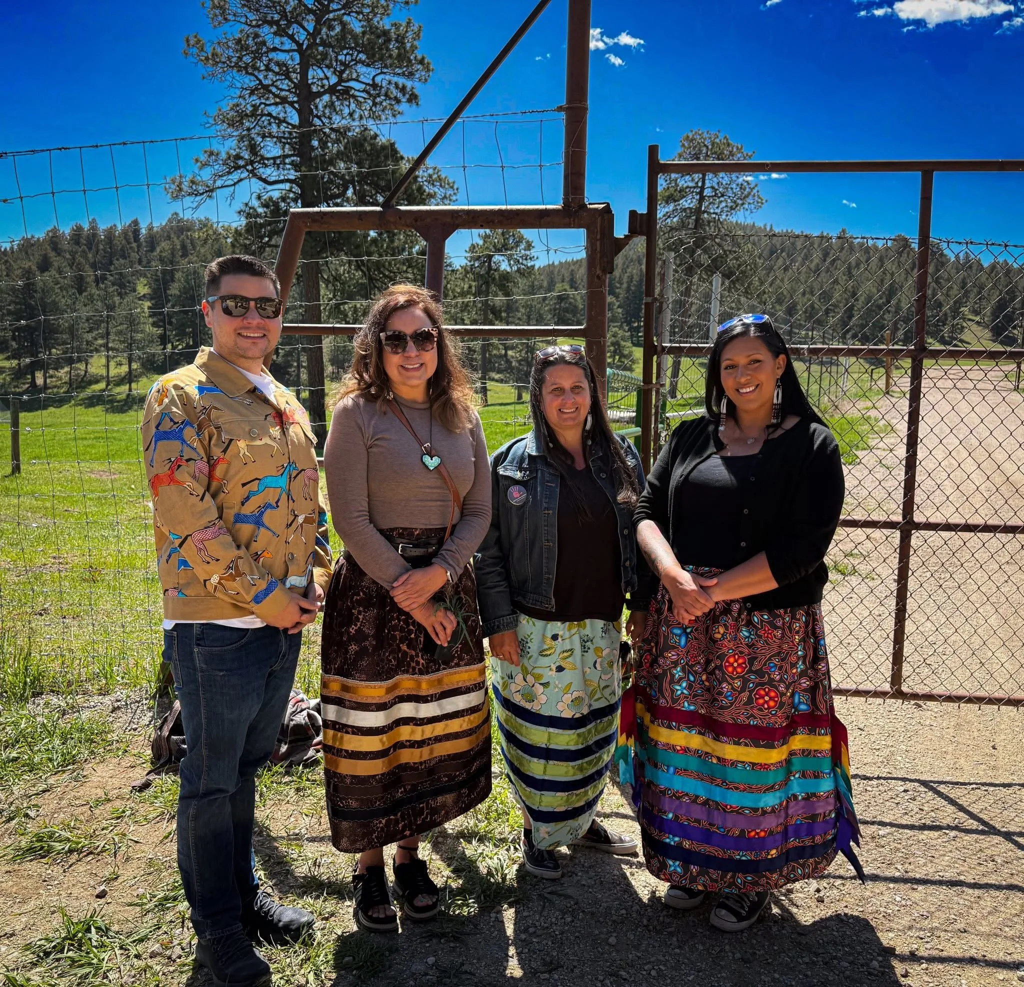 Four people standing outside near a chain-link fence, with trees and grass in the background, on a bright, sunny day.