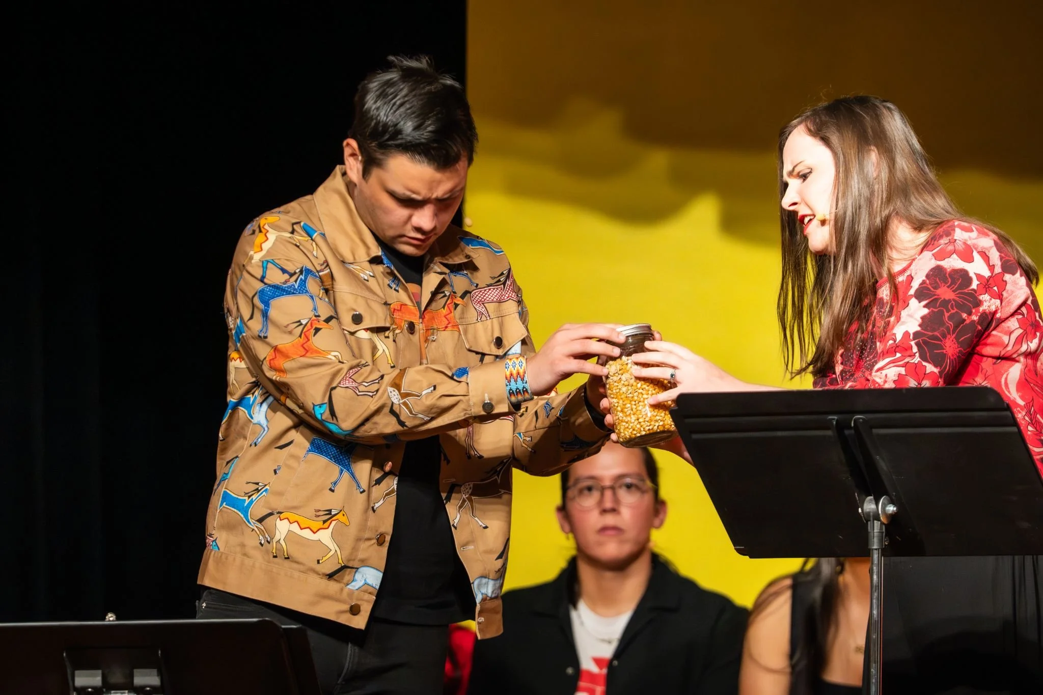 Two performers on stage exchange a jar of popcorn, with a woman in a red floral blouse and a man in a colorful horse-patterned jacket, while a person in glasses watches in the background.