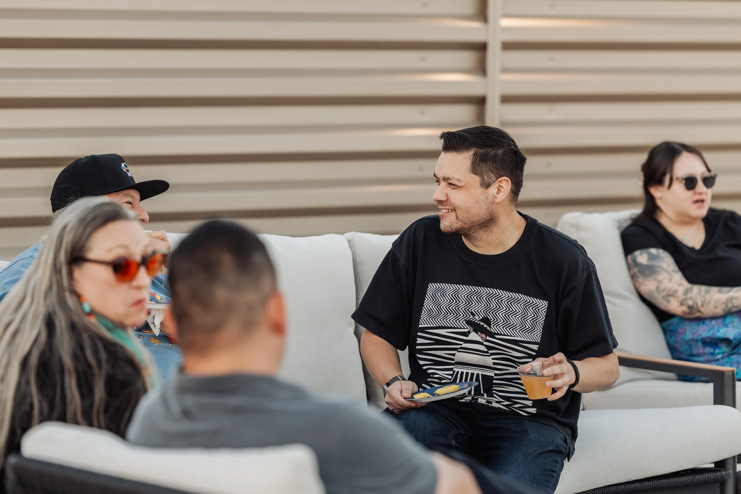 A group of people sitting on a outdoor sofa at a social gathering, with a young man smiling and holding a drink and plate of food, engaged in conversation, while others sit around him in a casual setting.