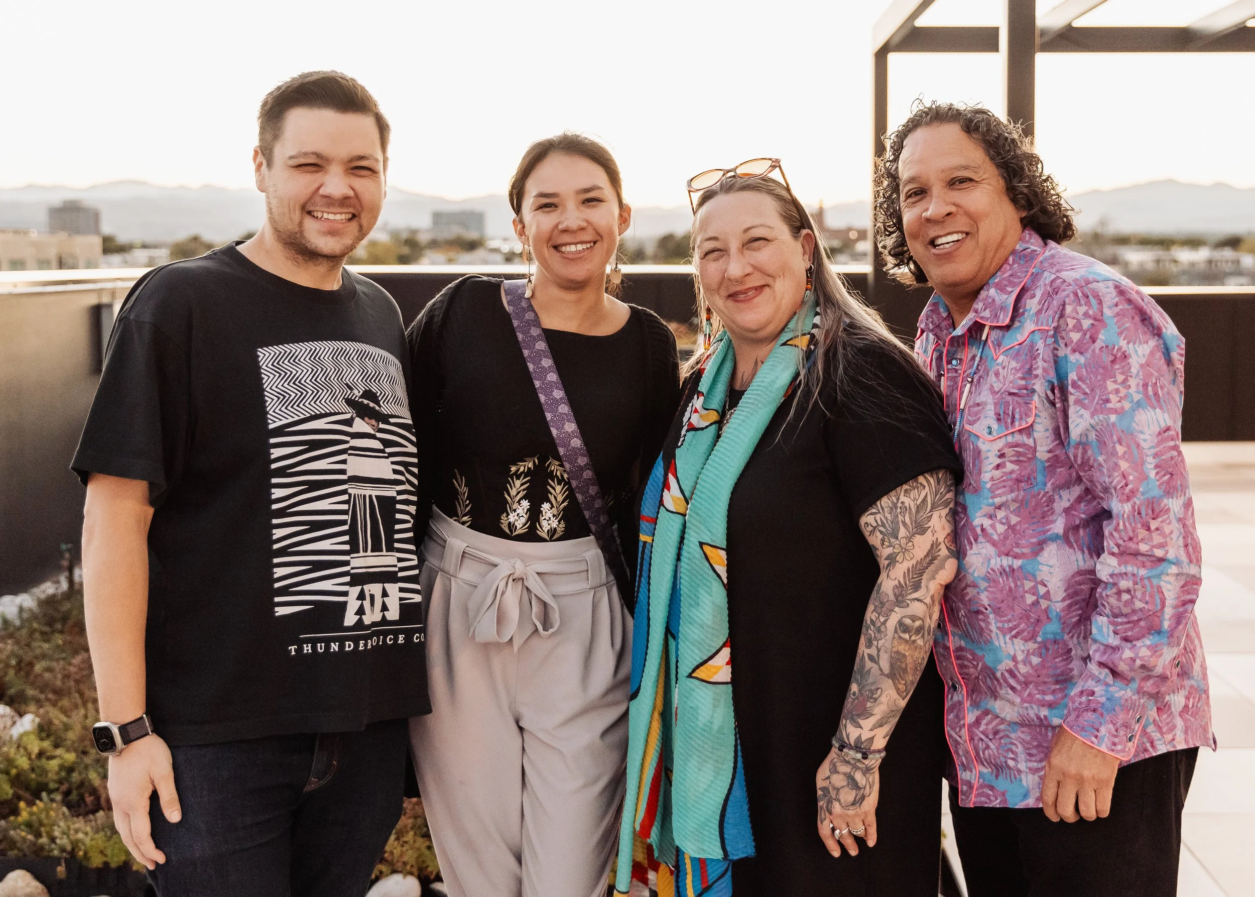 Four people standing outdoors on a rooftop, smiling at the camera with city and mountains in the background during sunset.