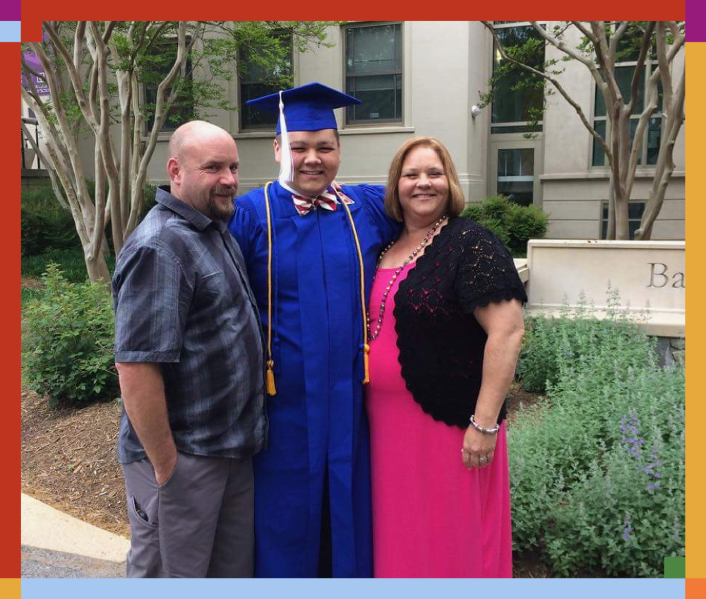 A young man in a blue graduation cap and gown with a red, white, and blue bow tie, standing between a man in a gray shirt and a woman in a pink dress with a black lace cover-up, all smiling outdoors.